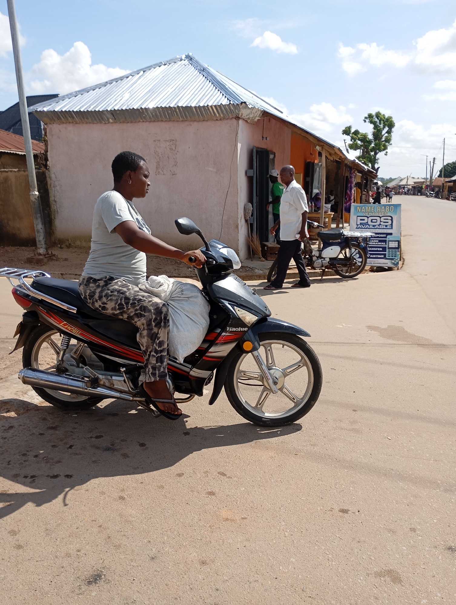 In Shendam, Nigeria, a woman is seated on a motorcycle, her hands on the handlebars. She is wearing a light grey t-shirt and patterned leggings, with dark flip-flops on her feet. The motorcycle is black and red with silver accents, and a large white bag is secured behind the seat. The woman is positioned on the right side of the motorcycle, facing away from the camera.
The scene unfolds on a paved road, with a building in the background that has a corrugated metal roof. To the right of the building, a man in a white shirt and dark pants walks away from the camera. A blue and white scooter is parked near the building. A sign advertising "NAME RABO VENTURES POS AVAILABLE" is also visible near the building. The sky above is blue with scattered white clouds. The environment appears to be an urban or semi-urban setting in Nigeria.