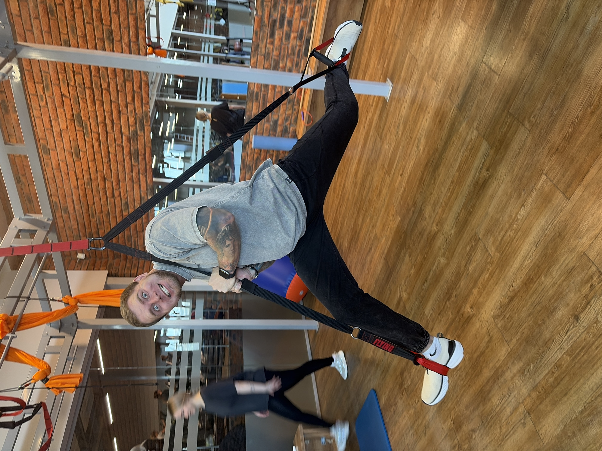 A medium shot depicts a man in a gym using suspension straps. The man is suspended upside down with his feet in the straps, wearing black pants, a grey shirt, and white shoes. He has light skin, short blond hair, and is looking directly at the camera with a slightly startled expression. His left arm is visible and has tattoos.
The gym setting includes a wood-patterned floor, brick and metal structures in the background, and other gym equipment. Other people are visible in the background of the gym, including a woman wearing a black top and black leggings and white shoes. The straps the man is using have the word "FLYING" printed on them.
The image was likely taken in Chaikovsky, Russia, based on contextual information provided.