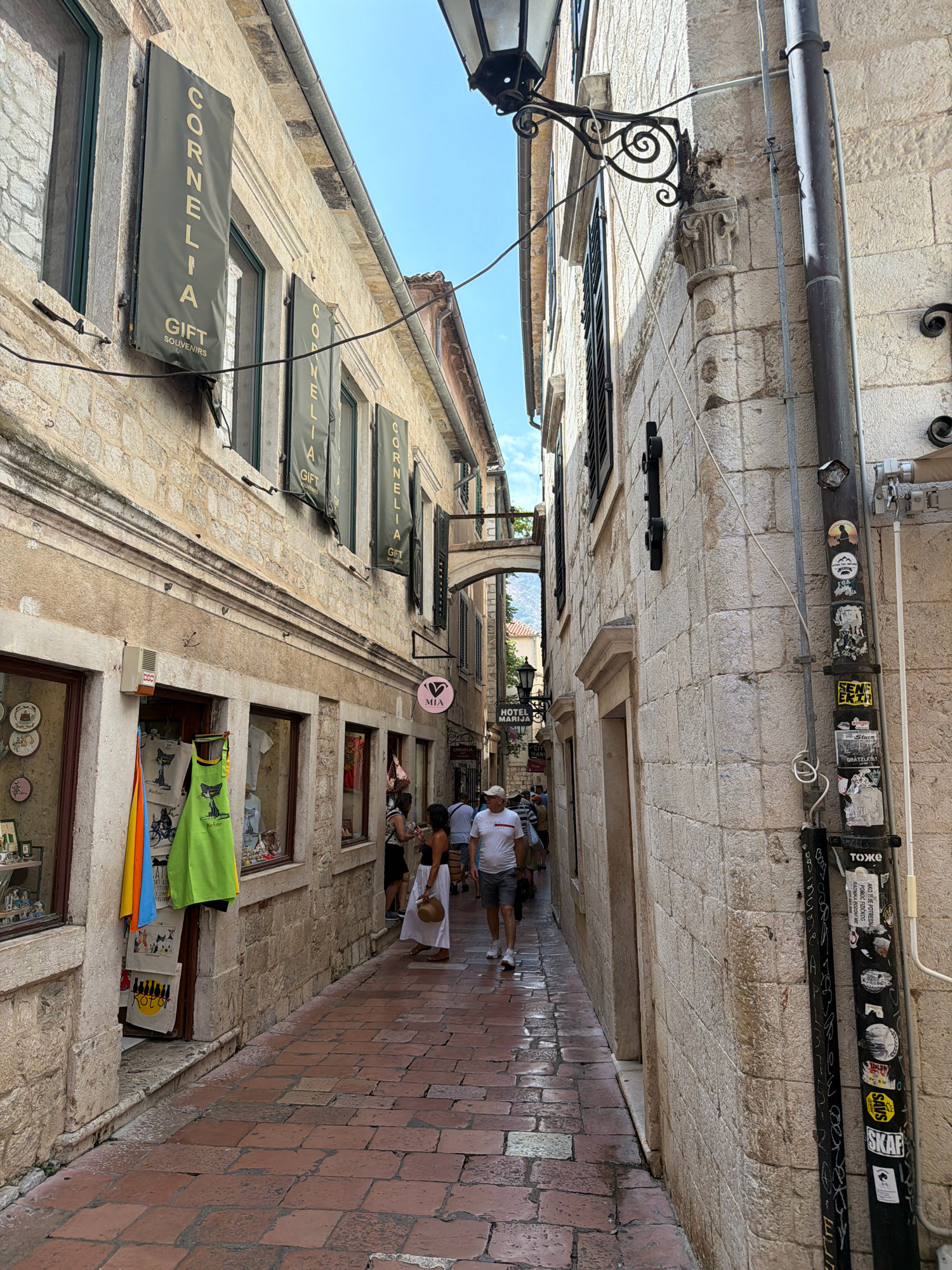 The image shows a narrow, cobblestone street in Kotor, Montenegro, lined with old stone buildings. On the left, a shop displays colorful textiles and souvenirs in its window. Above the shop, banners hang from the building, one reading "CORNELIA GIFT SOUVENIRS." On the right, a thick pipe runs vertically up the stone wall, covered in various stickers. A man wearing a white t-shirt and grey shorts walks down the street, away from the camera. Further down the street, several other people are visible, appearing to browse shops or walk. The architecture suggests a historic European town, consistent with Kotor's old town. The sky is blue with minimal clouds.