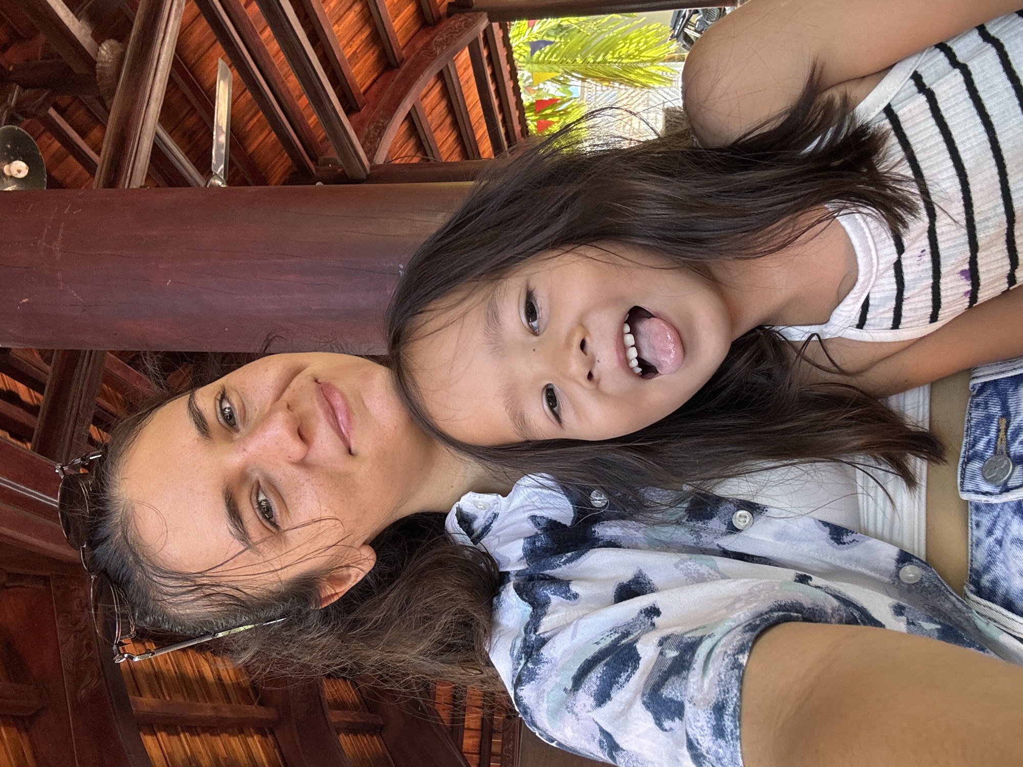 A woman and a young girl are pictured in a selfie. The woman, with light skin and dark hair, is wearing a patterned button-up shirt with a white under-shirt and a denim jacket, while the girl, with dark hair and light skin, is wearing a white and black striped top. The girl has her tongue sticking out. They appear to be under a wooden structure with exposed rafters. The location is Mũi Né, Vietnam.