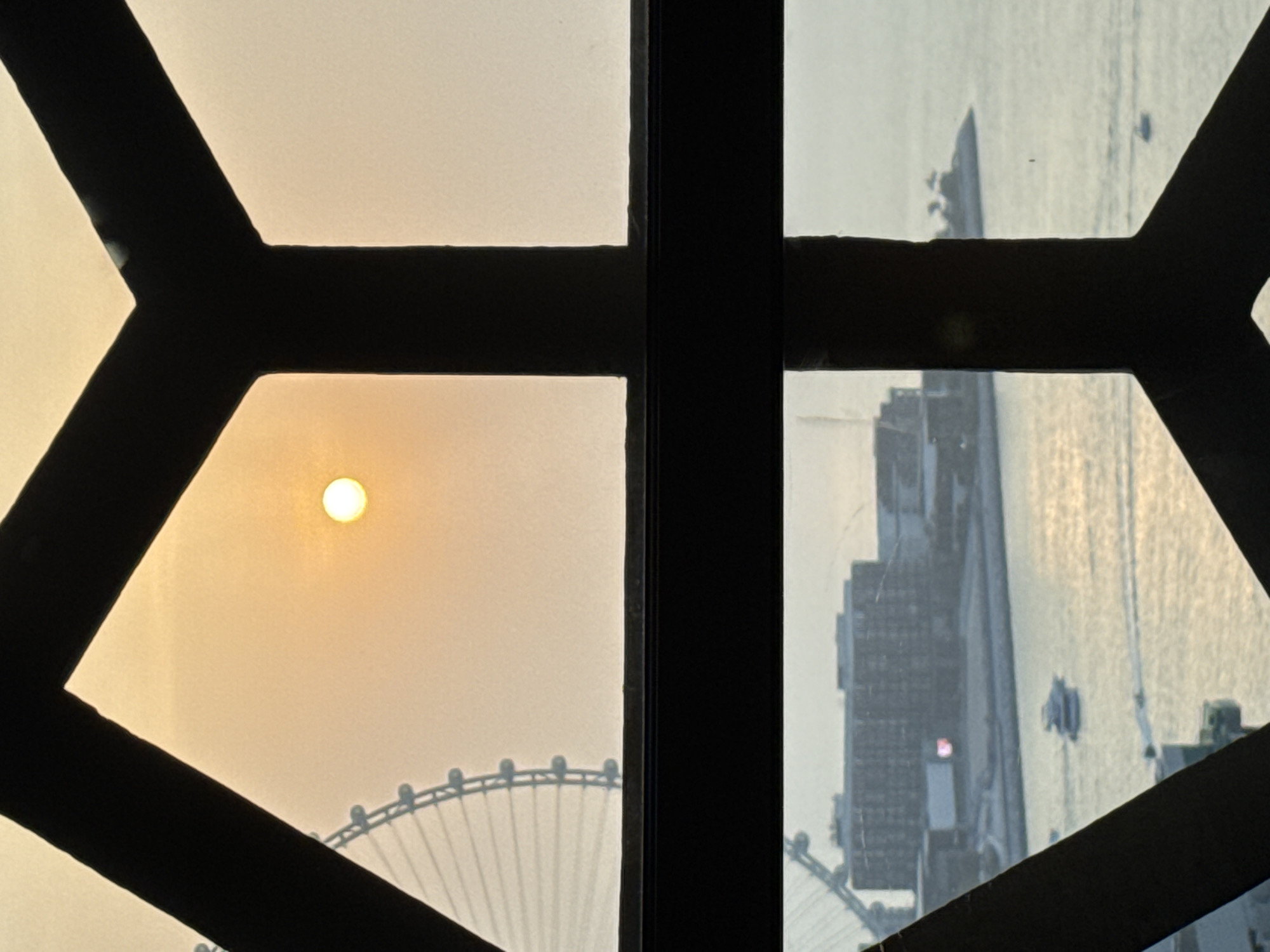 The image captures a view from a window with dark framing, divided horizontally by a thick black bar. The upper portion of the image shows a hazy, orange-tinted sky with the sun as a bright, small orb. To the left, a large Ferris wheel, possibly the Ain Dubai, is visible in silhouette against the sky. The lower portion of the image depicts a body of water, reflecting the soft light. On the water, several boats are navigating. In the background, there is a large building complex with a modern architectural style. The scene suggests an urban waterfront setting in Dubai, United Arab Emirates, likely during sunrise or sunset due to the warm lighting and hazy atmosphere.