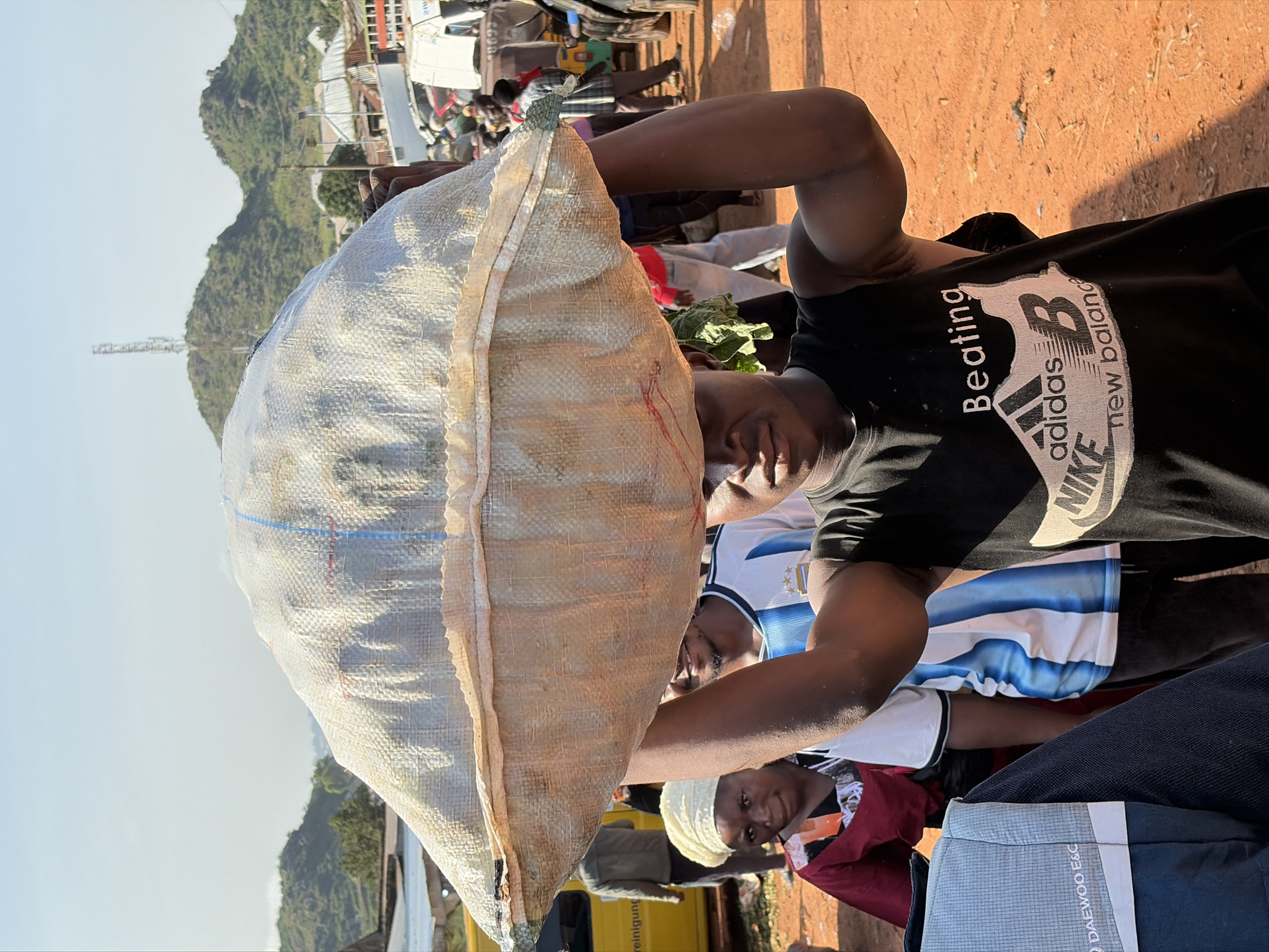 In Gura Top, Nigeria, a young man carries a large, woven sack filled with goods on his head. The sack is white with blue and red stripes. He wears a black t-shirt with multiple brand logos, including Adidas, Nike, and New Balance. Behind him, a woman in a red and white headscarf looks to the left, and a man in a blue and white striped soccer jersey stands partially obscured. The background shows a dusty street with vehicles, including a yellow truck on the left and various other cars and a tuk-tuk further back. In the distance, green hills rise under a clear, bright sky, with a cell tower visible on one of the peaks. The ground is a reddish-brown dirt.