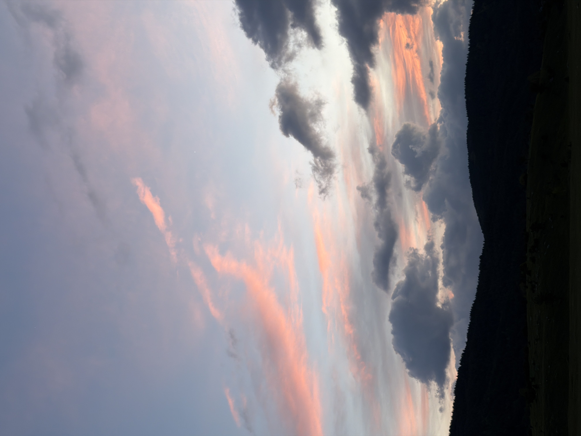 The image depicts a twilight sky in Njegovuđa, Montenegro. The sky transitions from a light blue in the lower left to shades of pink and orange near the horizon. Wispy, pink-tinged clouds are scattered throughout the left portion of the frame. Along the right edge, darker, gray cumulus clouds are present. A dark, heavily forested mountain range runs along the right side of the image, silhouetted against the colorful sky. A strip of green field is visible at the very bottom right edge.