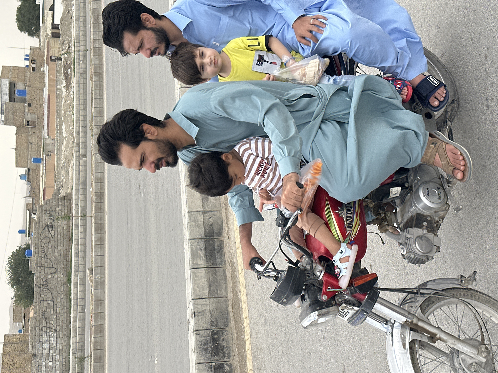 On an urban street in Quetta City Tehsil, Pakistan, a red motorcycle is oriented from the lower right to the upper left. Three individuals are on the motorcycle, two adults and a child in the middle of the bike. All three people wear traditional clothing, in hues of light blues and yellows. The motorcycle rider is wearing blue pants and tan sandals. The motorcycle has some visible dirt and is in an area with paved roads. To the left, along the roadside, is a concrete barrier. Beyond that is a residential area with stone buildings.