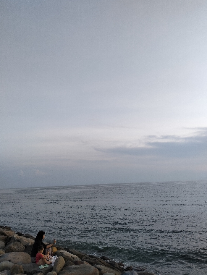 A woman wearing a hijab and a child are sitting on rocks at the edge of a body of water. The water is choppy with small waves. The sky is overcast, and a thin line of clouds is visible on the horizon. The rocks are large and varied in shape and size. The woman is looking out at the water, and the child is sitting beside her, also appearing to gaze at the sea.