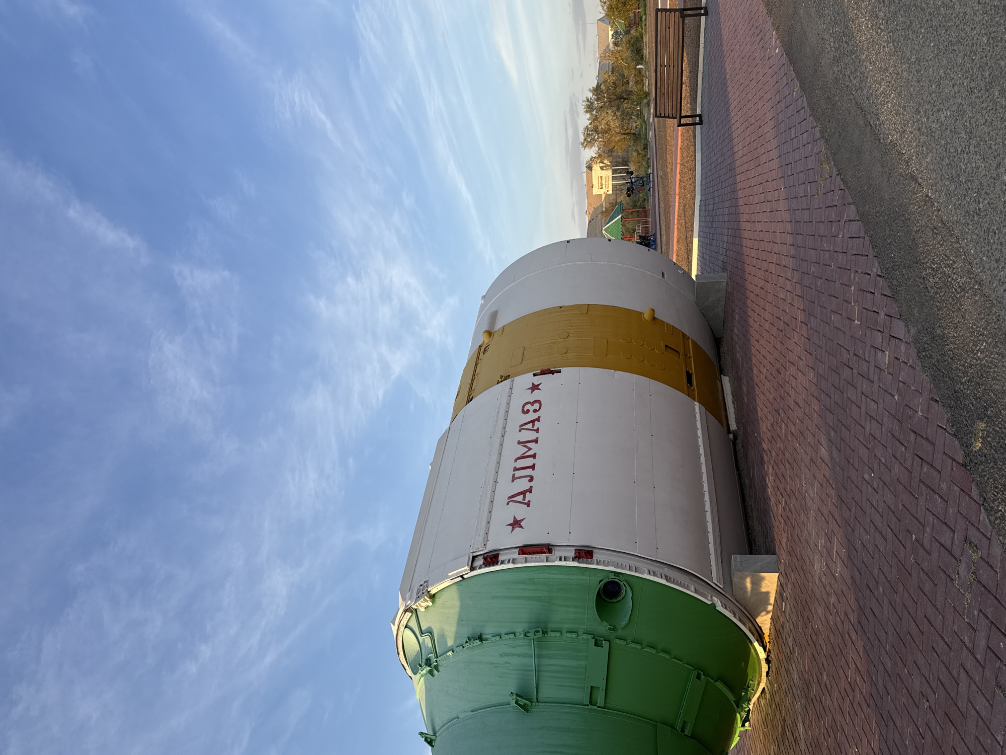 An outdoor, low-angle, full shot shows a section of a large, cylindrical object painted green, white, and yellow, with red lettering that reads "АЛИМАЗ" flanked by two red stars. The object is resting on a concrete support and appears to be a decommissioned spacecraft or rocket booster. It is positioned on a red brick paver walkway, with a dark asphalt path in the foreground. In the background, there is a small park area with playground equipment and a wooden bench. Beyond the park, low-rise buildings and trees are visible under a bright blue sky with wispy white clouds. The scene is located in the city of Baikonur, Kazakhstan, a site historically associated with space exploration.