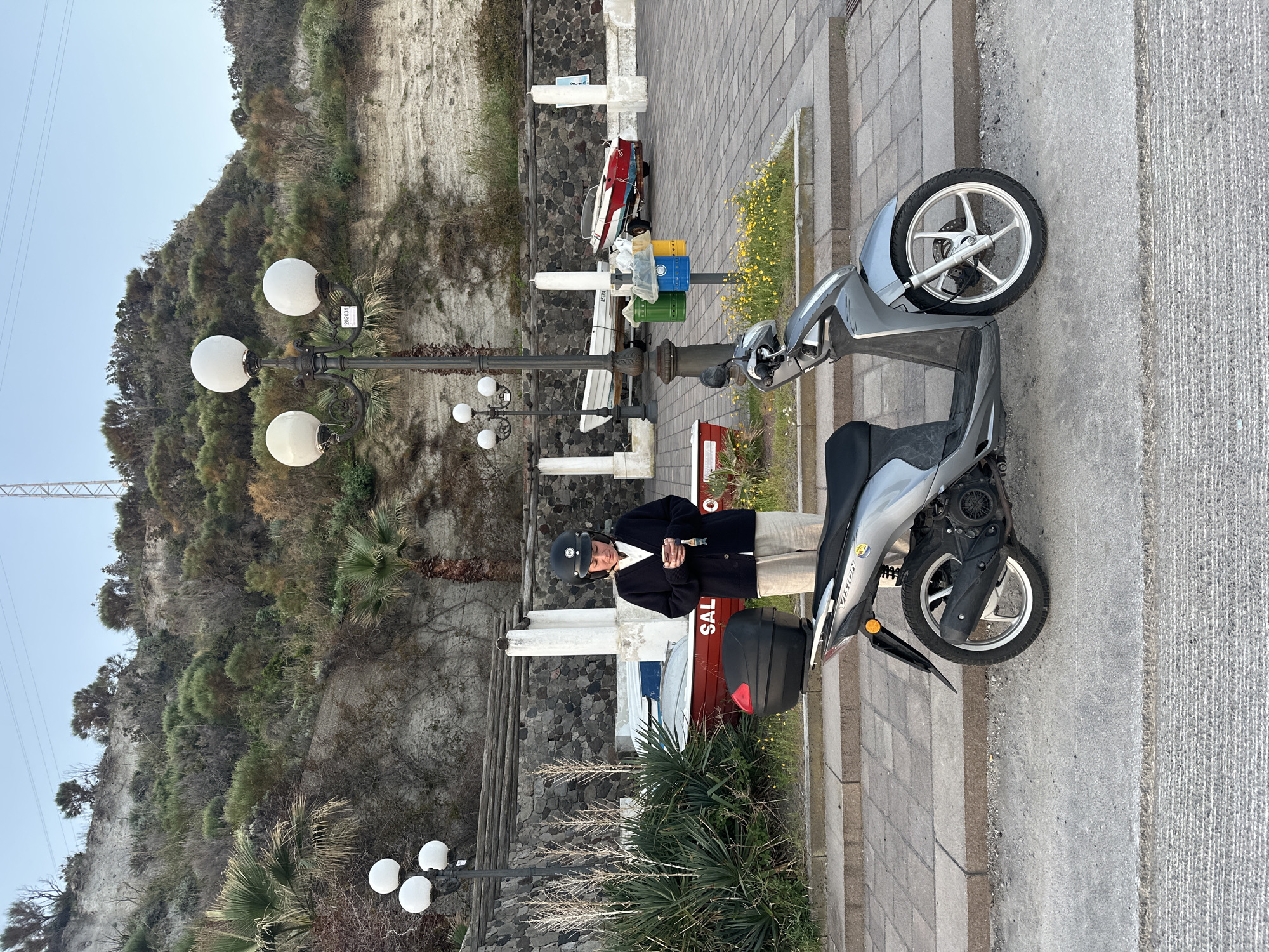 The image captures a person standing next to a silver scooter on a paved walkway in what appears to be Lipari, Italy. The individual, wearing a black helmet, a dark jacket, and light trousers, is seen holding a small item in their hands, possibly preparing for or just finishing a ride. The scooter is light grey with a black seat and a black storage box at the rear, displaying the word "VISION" on its side panel.
Behind the person and scooter, a tall, steep cliff covered with sparse, dry-looking vegetation rises under a clear sky. The ground consists of large, light-colored paving stones, with a darker stone wall separating it from a section of unkempt earth and shrubbery where small yellow flowers grow. Several vintage-style lampposts, each featuring multiple spherical white lights, are positioned along the path. One lamppost has a white sticker with the number "282035".
Further into the background, beyond the lampposts, are three colored trash bins (green, blue, and yellow) and two small boats. One boat is predominantly red and white, with the partial word "SAL" visible on its side, while the other appears white. Palm trees and other resilient Mediterranean plants are scattered across the landscape. The lighting suggests it is daytime, possibly morning or late afternoon, with soft, even illumination. The scene conveys a relaxed coastal atmosphere.