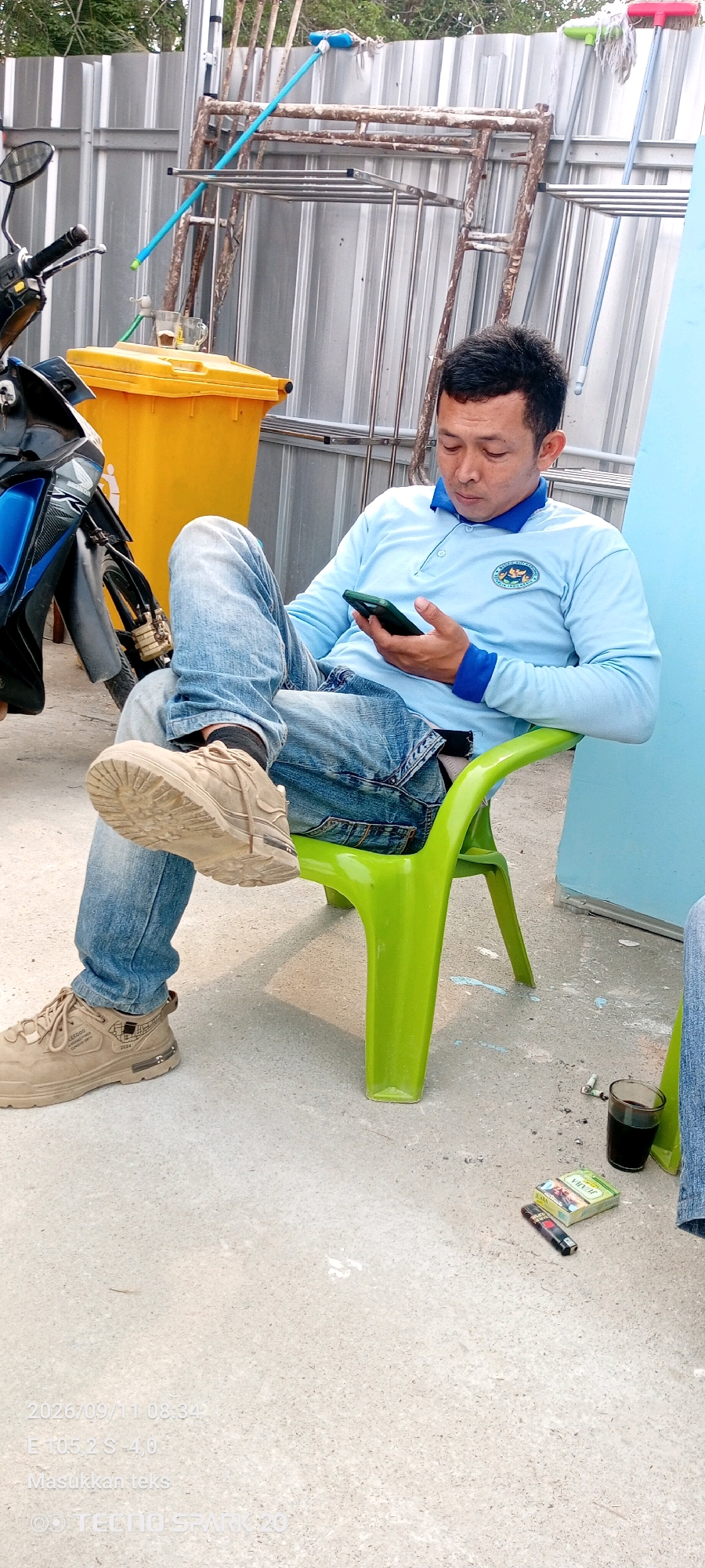 A man wearing a light blue long-sleeved shirt and blue jeans is seated on a green plastic chair. He is holding a green smartphone in his right hand, looking down at it. His left leg is crossed over his right. A motorcycle is partially visible to the left of the frame, with a yellow trash bin behind it. In the background, there is a corrugated metal fence and some metal scaffolding. On the ground near the chair, there is a pack of cigarettes, a lighter, and a glass of dark liquid. The ground is concrete. The timestamp overlay indicates the image was taken on 2026/09/11 at 08:34. The text overlay "Masukkan teks" and "TECNO SPARK 20" suggest the image was taken with a TECNO SPARK 20 phone and has an editable text element. The scene appears to be outdoors in an urban or semi-urban environment, possibly in Simpang Pematang, Indonesia, given the timestamp and text overlay.