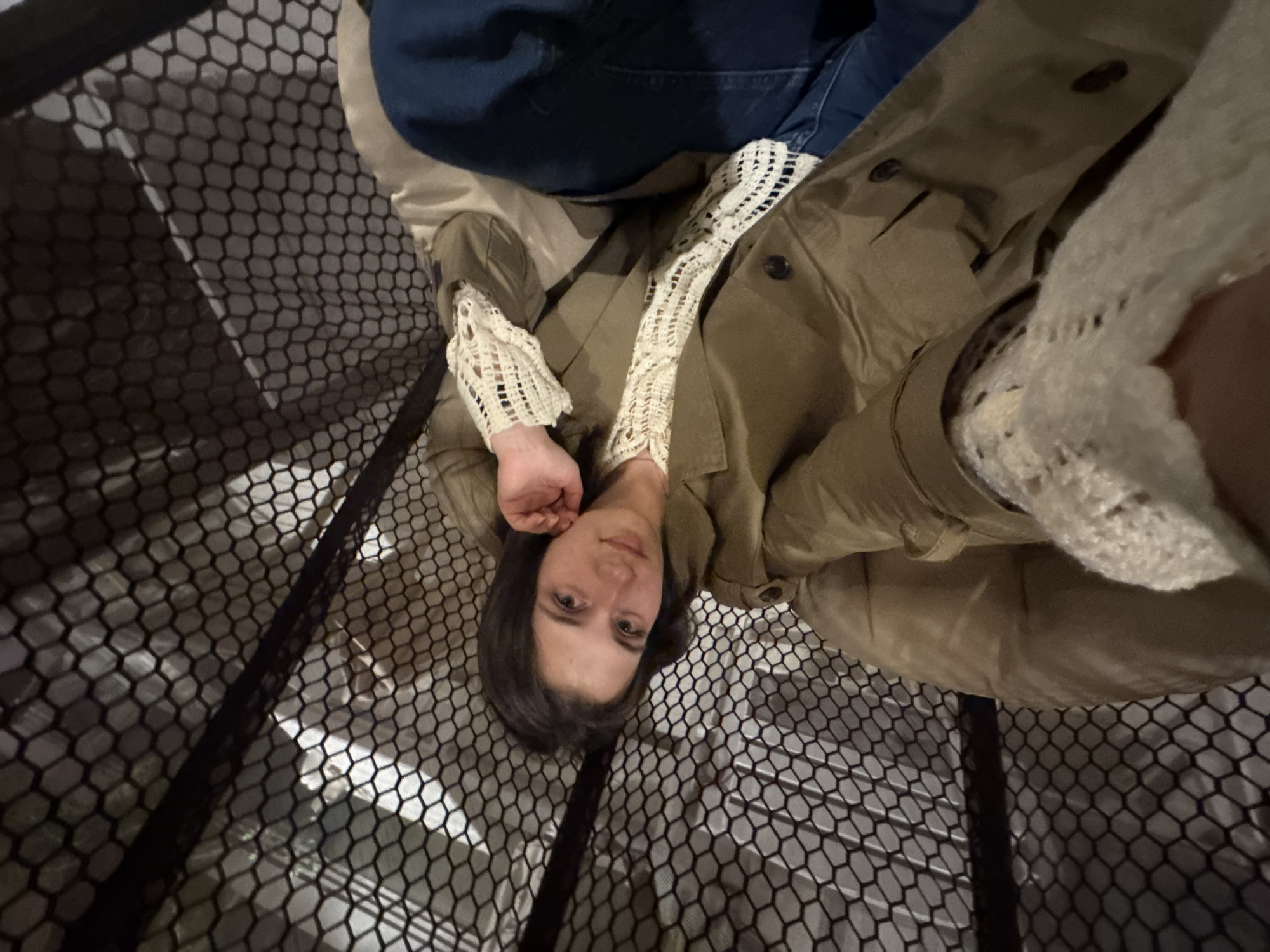 An angled, high-perspective photograph shows a young woman resting within a structure featuring a prominent black hexagonal mesh pattern. The mesh dominates the foreground and extends into the background, partially obscuring the environment behind it. The woman has dark hair and light eyes, looking directly at the camera with a neutral, calm expression. She is wearing a dark olive or khaki-colored jacket or coat with visible buttons. Her arms are adorned with distinctive cream-colored, open-weave crocheted or knitted sleeves/cuffs. A portion of dark blue fabric, likely denim jeans, is visible towards the upper part of the frame. Her head rests on a light beige surface, possibly upholstery or clothing. The background, seen through the mesh, appears to be a light-colored, possibly tiled or patterned floor or wall, with some darker areas that could be shadows or structural elements. The setting suggests an indoor or enclosed architectural space, possibly a public building, museum, or an area with a unique protective or decorative mesh barrier. No text is visible. The scene takes place in Perm, Russia.