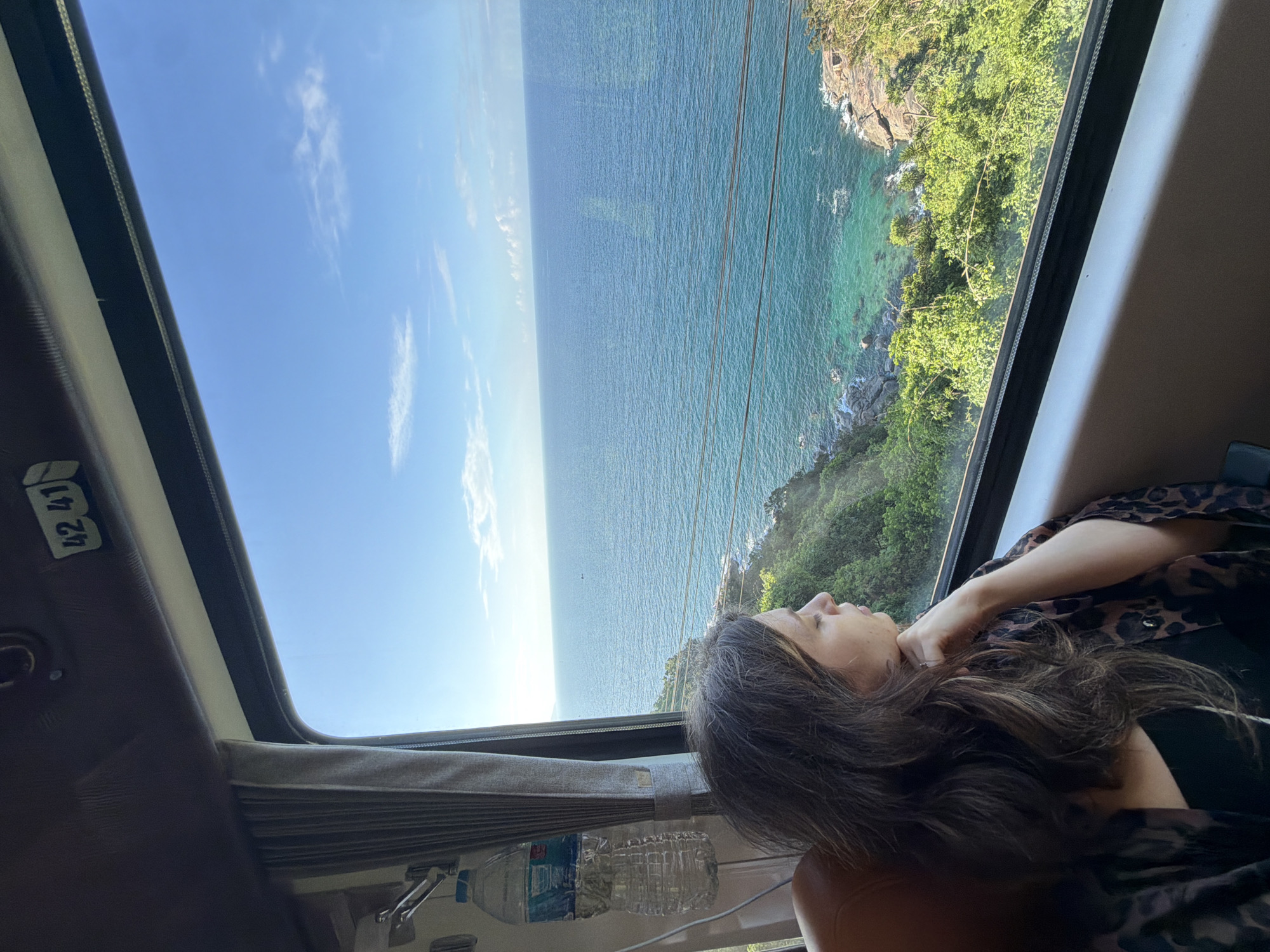 A medium shot depicts a woman inside a moving vehicle looking out a window. The vehicle is likely a train or a bus, based on the interior paneling and window size. The window provides a scenic view of the ocean and a rocky coastline covered in green vegetation. The sky is blue with a few clouds. The woman has long, dark hair and is wearing a patterned shirt. A water bottle sits near the window. A sticker with the numbers "42 41" is visible on the vehicle’s interior. While the image features an ocean view, there is no immediate indication that this scene is in Huế, Vietnam. Further visual or contextual information would be needed to confirm the location.