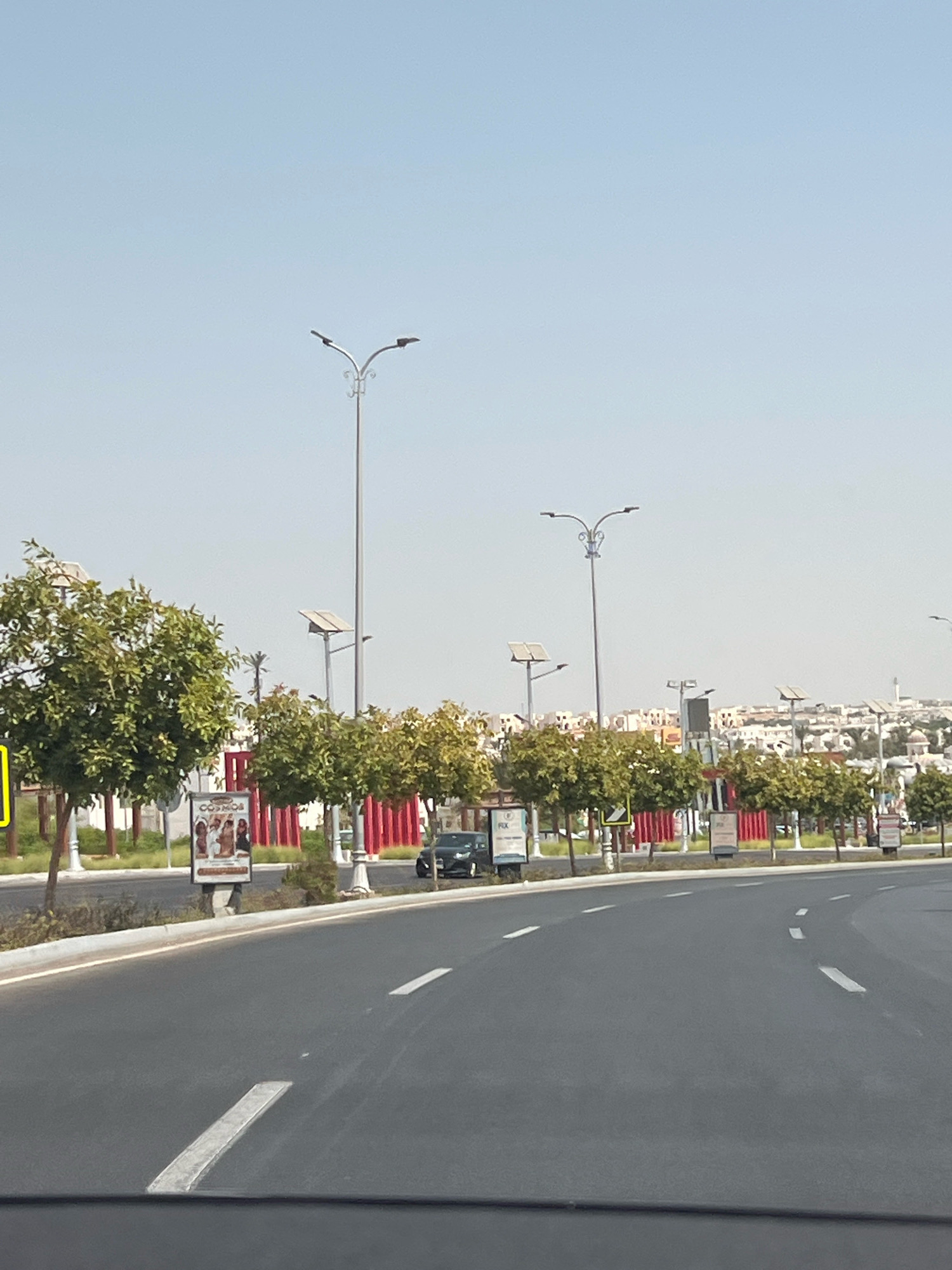 The image depicts a wide, paved road curving to the right, characteristic of a modern urban development in Sharm El Sheikh, Egypt. The road is marked with white dashed lines and a solid white line, indicating lane divisions. On the left side of the road, a row of neatly planted trees with lush green foliage lines a sidewalk. Behind the trees, a series of billboards or advertisements are visible, one prominently displaying the word "COSMOS". A dark-colored car is visible driving on the road. Further in the background, more trees and buildings, some with distinctive architectural elements such as domes, suggest a developed residential or commercial area. Several tall streetlights, some equipped with solar panels, are strategically placed along the roadside, illuminating the urban landscape. The sky is clear and pale blue, indicating a sunny day. The overall impression is one of a well-maintained and modern cityscape, likely a newer section of Sharm El Sheikh designed for vehicular traffic and pedestrian accessibility.