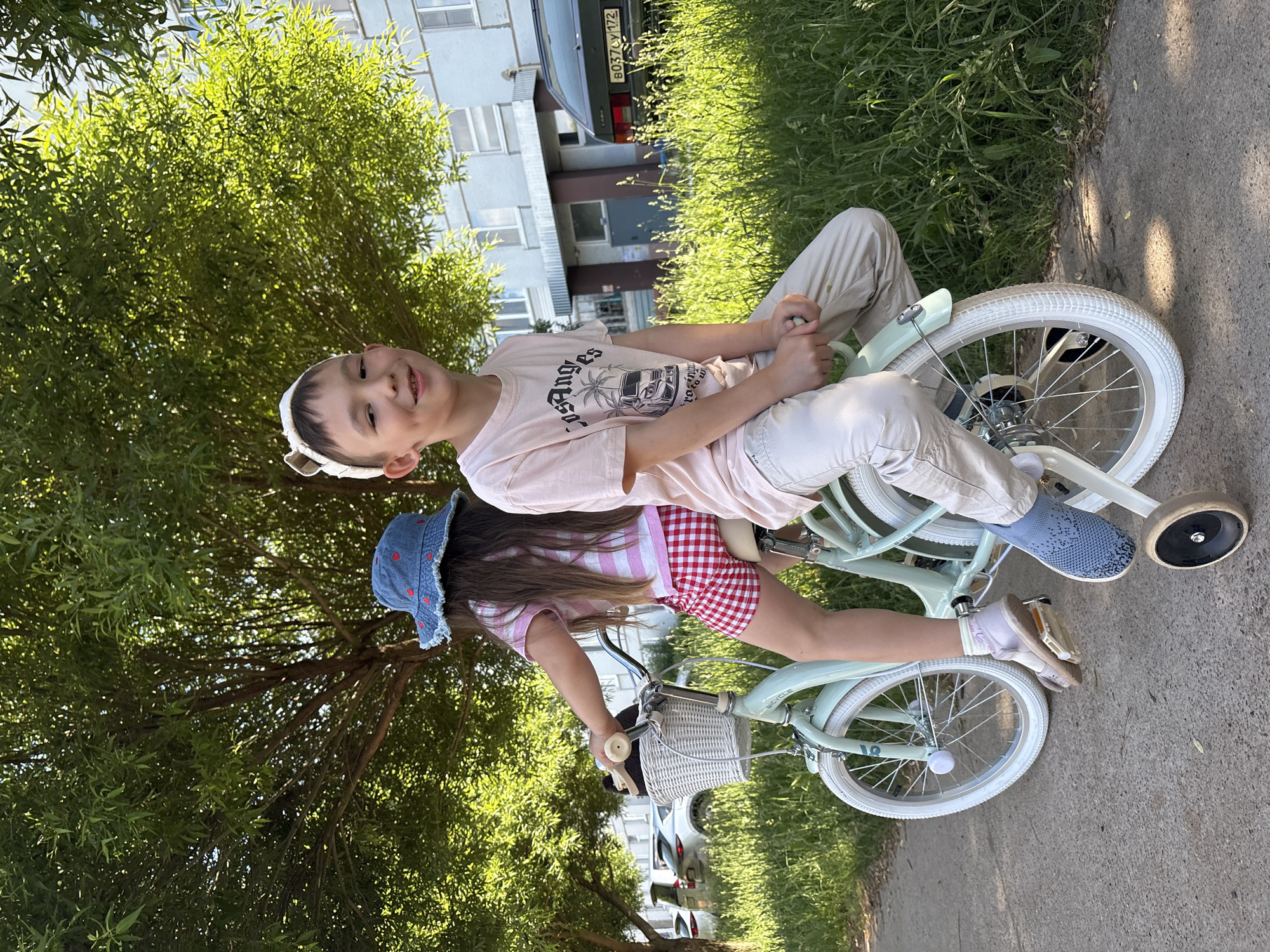 A boy and a girl are depicted sharing a light mint green children's bicycle equipped with white training wheels, positioned on a grey paved path under bright daylight in Chaikovsky, Perm Krai, Russia. The boy, exhibiting light skin and dark hair, wears a light-colored headband with ear-like protrusions, a light pink T-shirt featuring a "Los Angeles" graphic, light beige pants, and light blue slip-on shoes. He is seated on the rear fender of the bicycle, facing forward and smiling towards the camera. The girl, with long dark hair, wears a blue denim bucket hat adorned with red heart patterns, a pink and white striped T-shirt, and red and white gingham shorts, with light pink shoes and white socks. She occupies the bicycle's main seat, her back mostly to the camera, with her feet on the pedals. The bicycle has a white woven basket attached to the handlebars and white-walled tires.
The immediate environment includes a strip of green grass bordering the paved path, with a large, leafy tree providing shade and dominating the upper background. In the background, a light-colored, multi-story residential building with multiple windows is visible. To the right of the building, a dark green VAZ-2107 series car with Russian license plate "B037CY 172" is parked, partially obscured by grass. Further down the path to the left, several other vehicles, including at least two white SUVs, are parked.