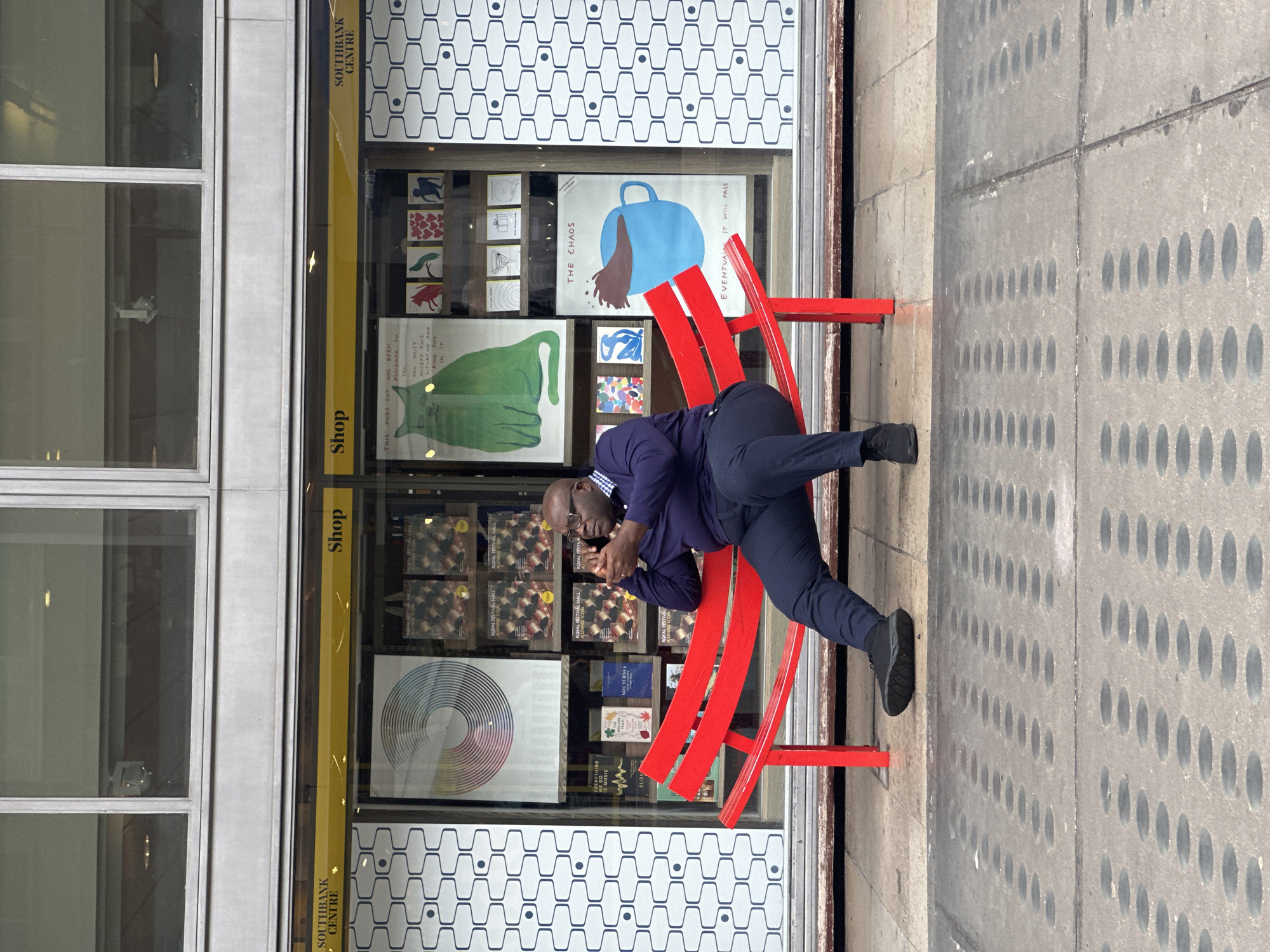 A man wearing glasses, a dark blue shirt, and dark blue trousers sits on a bright red bench in front of a shop window. The shop is the Southbank Centre shop in London, United Kingdom. The shop's display includes posters with artistic designs, books, and vinyl records. The man is leaning forward, looking at his hands, and appears to be resting. The background features the exterior of a building on the left and a sidewalk with a textured surface on the right.