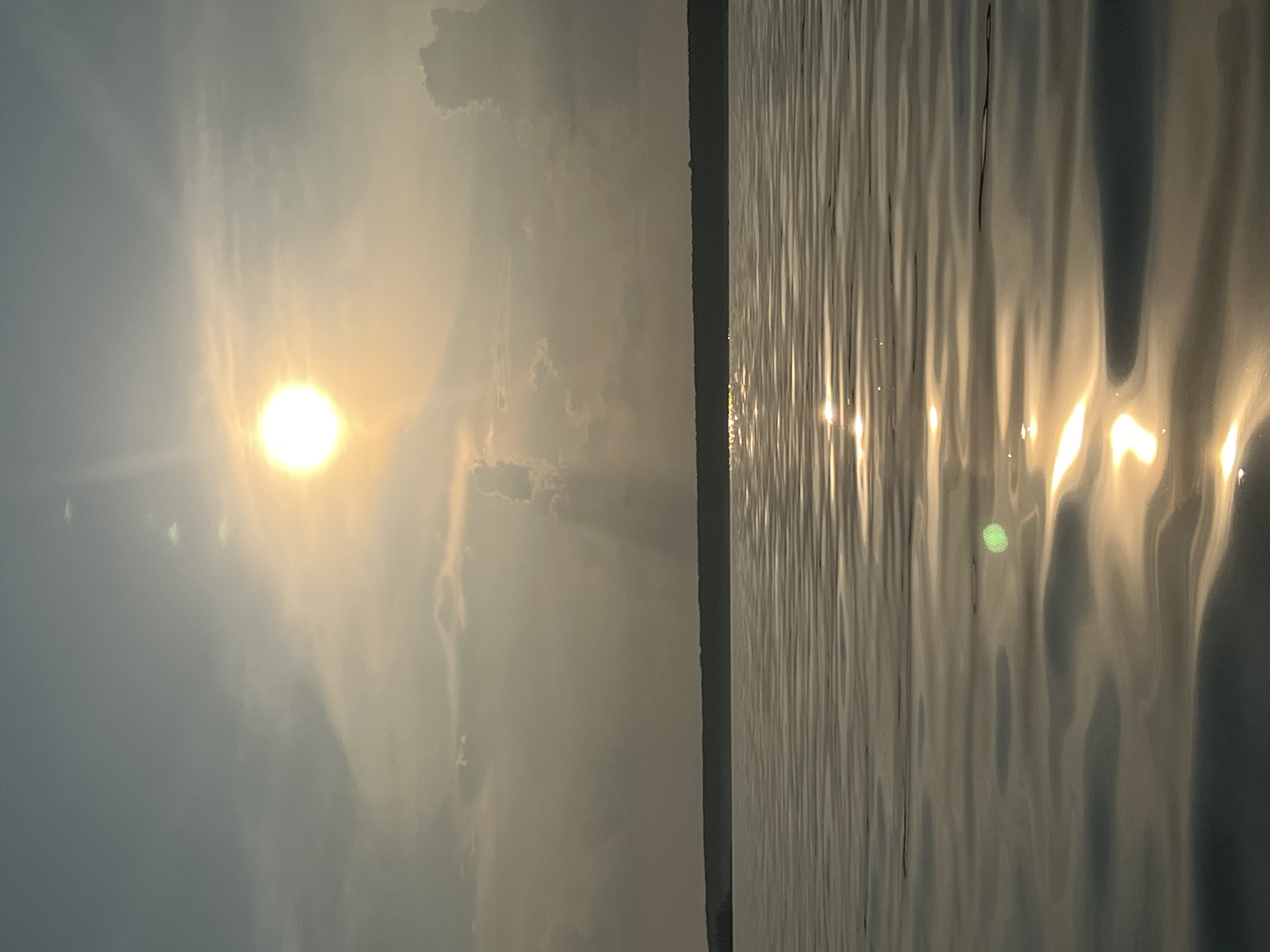 The image shows a body of water with a distant shoreline under a hazy, sunlit sky. The sun is high and bright, with rays of light filtering through the clouds. The water is calm with ripples reflecting the sunlight. The shoreline is a dark, silhouetted strip of land, possibly covered in trees. The overall impression is one of a serene, natural landscape, likely a lake or a large river. The location in Borovoe, Russia is not discernible from the image alone. There are no people or discernible objects in the image besides the natural elements.