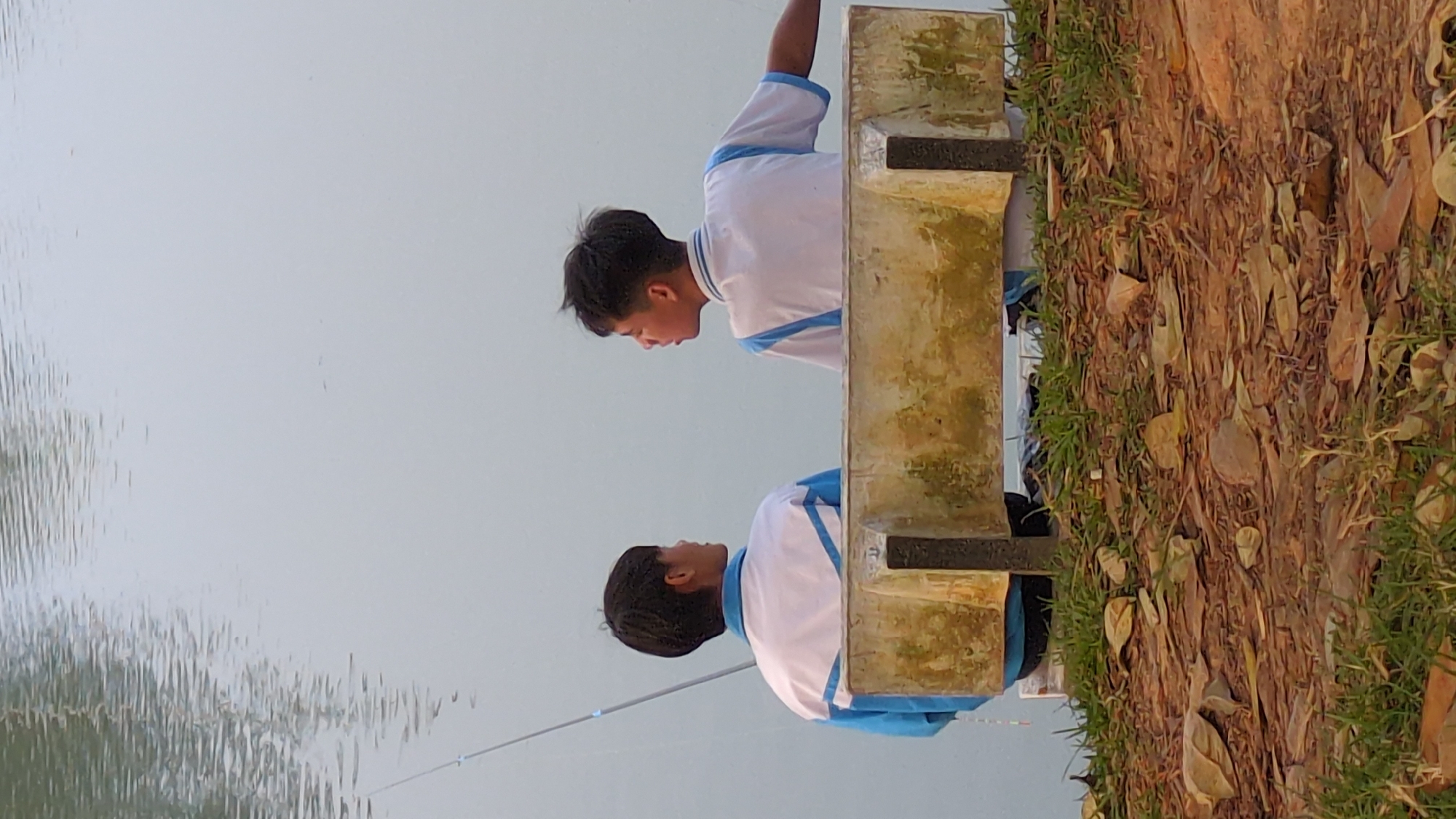 Two young men are fishing side-by-side, facing a body of water. They are wearing matching white shirts with blue trim. They are positioned near the edge of the water, which is bordered by dirt, grass, and fallen leaves. The location is Xuân Hương Lake in Đà Lạt, Vietnam. A cement structure separates the two men. Fishing poles extend from where they stand into the lake.