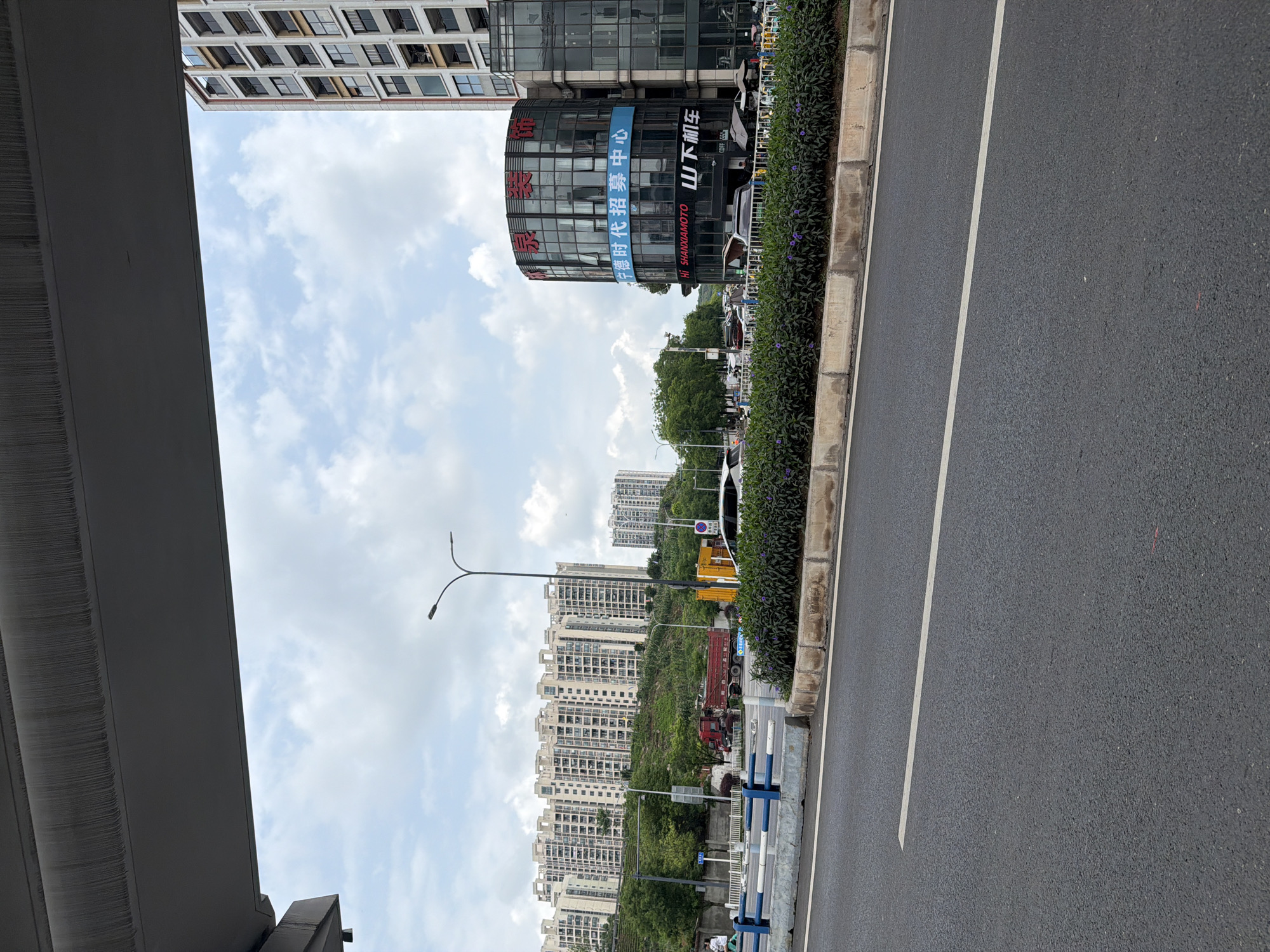 The image shows a street scene in Chongqing High-tech Zone, China. The view is of a multi-lane road with dense shrubs separating the sidewalk from the street. High-rise buildings are visible in the background. A building with commercial signage in Chinese characters is on the left. An overpass structure is visible on the left edge of the image. The sky is partly cloudy. There are several large trucks on the road.