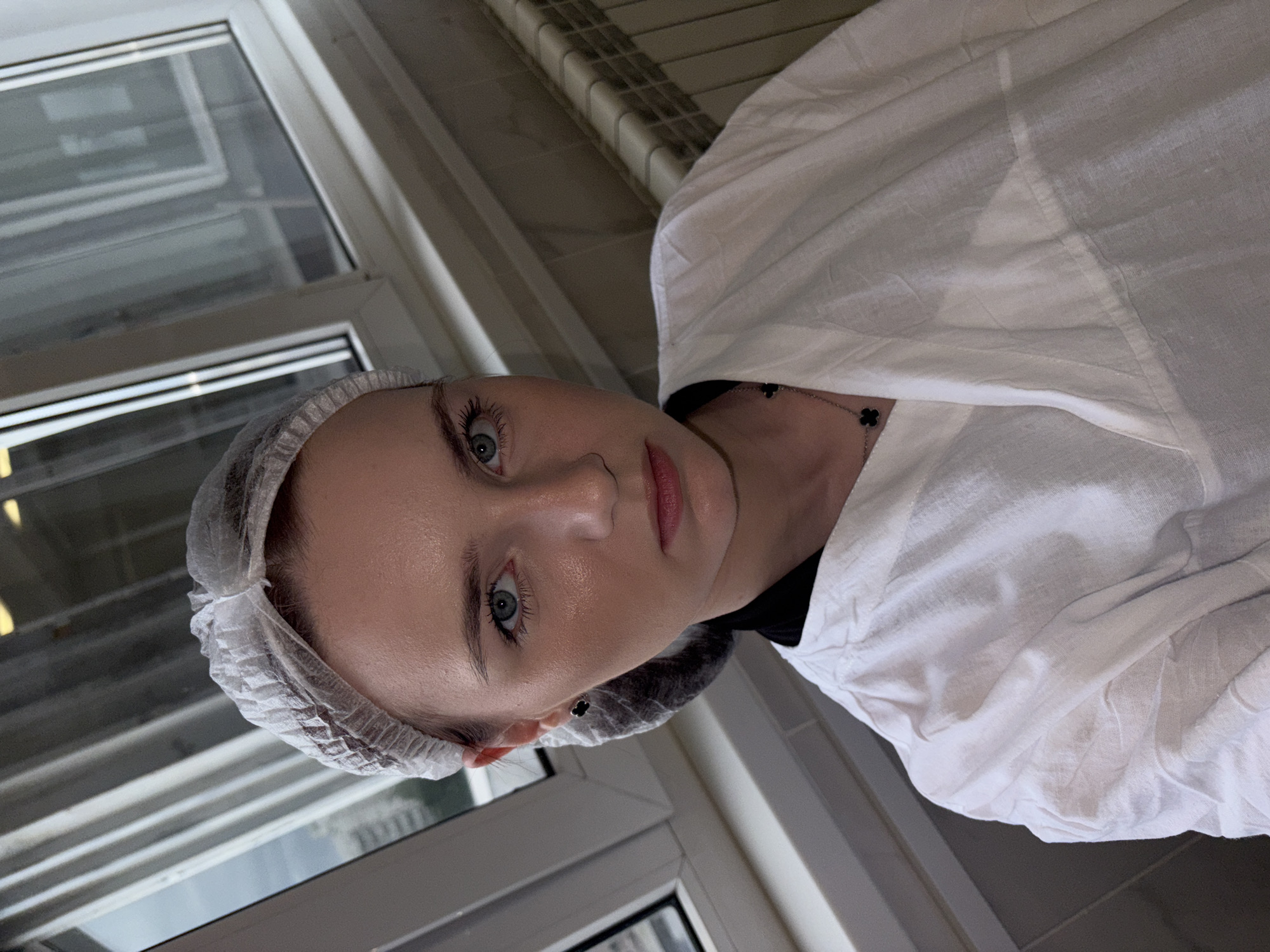 A young Caucasian woman is photographed from the chest up, looking directly at the camera. She is wearing a white disposable hair net and a white, V-neck garment that appears to be a lab coat or medical smock. A dark necklace with a clover-shaped pendant is visible on her chest. Her expression is neutral. The background includes a window with vertical blinds and a radiator. The lighting suggests an indoor setting. The location is given as pos. №13, Russia.