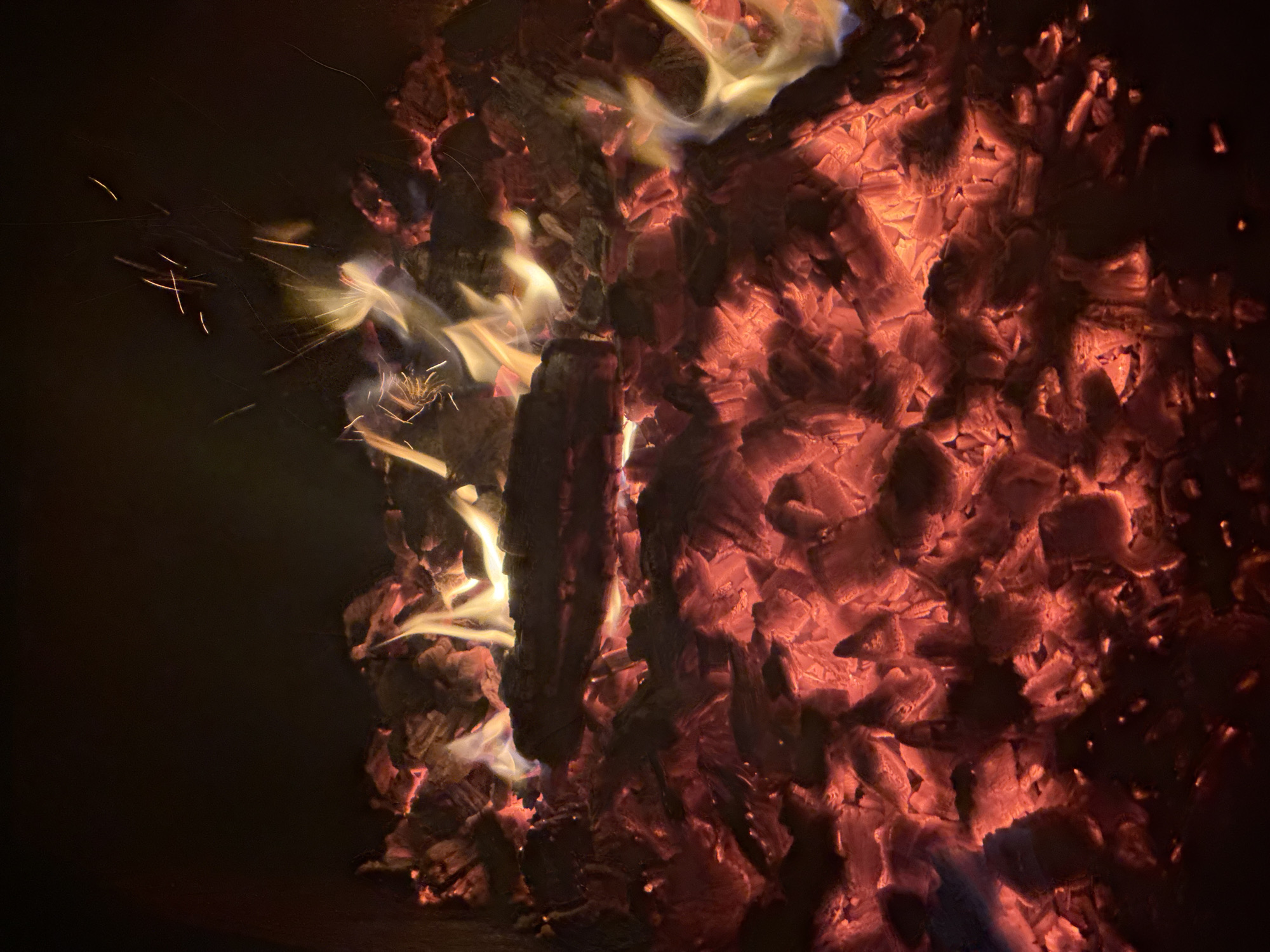 The image depicts a close-up view of glowing embers and burning wood. The scene is dark, with the primary light source coming from the intense orange and red glow of the fire. Several pieces of wood are visible, some partially burned and others engulfed in flames, casting dynamic yellow and white highlights. Small sparks are scattered throughout the upper portion of the image, indicating active combustion. The texture of the embers appears rough and uneven, with the flames creating a sense of movement and heat. The background is a deep black, suggesting a lack of ambient light and focusing all attention on the fire. There is no indication of any specific location within the city of Таганрогский, Russia, or any people present in the image.