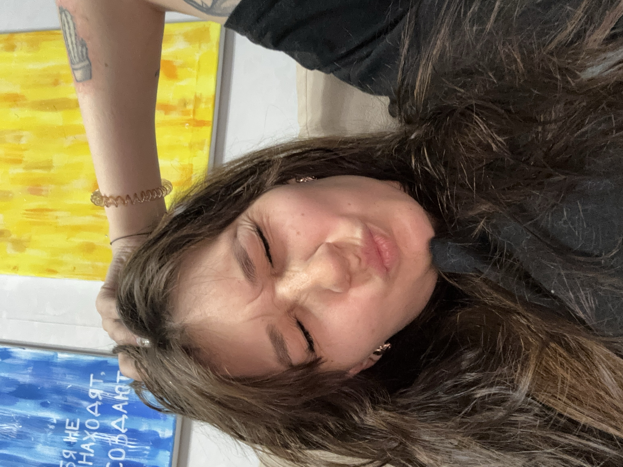 The image shows a close-up, selfie-style shot of a young woman with long, dark brown hair. Her eyes are closed, and her face is scrunched up in an exaggerated pout or grimace. She is wearing a black top and a small, circular earring in her left ear. Her right arm is raised and bent, with her hand on top of her head, revealing a tattoo on her forearm and a spiral bracelet on her wrist. In the background, there are two abstract paintings on a white wall, one predominantly blue with white text and the other yellow with streaks of orange. The woman appears to be indoors, possibly on a couch, given the beige upholstery visible on the right side of the frame. There is no identifiable information within the image that places the scene specifically in Novokuznetsk, Russia.