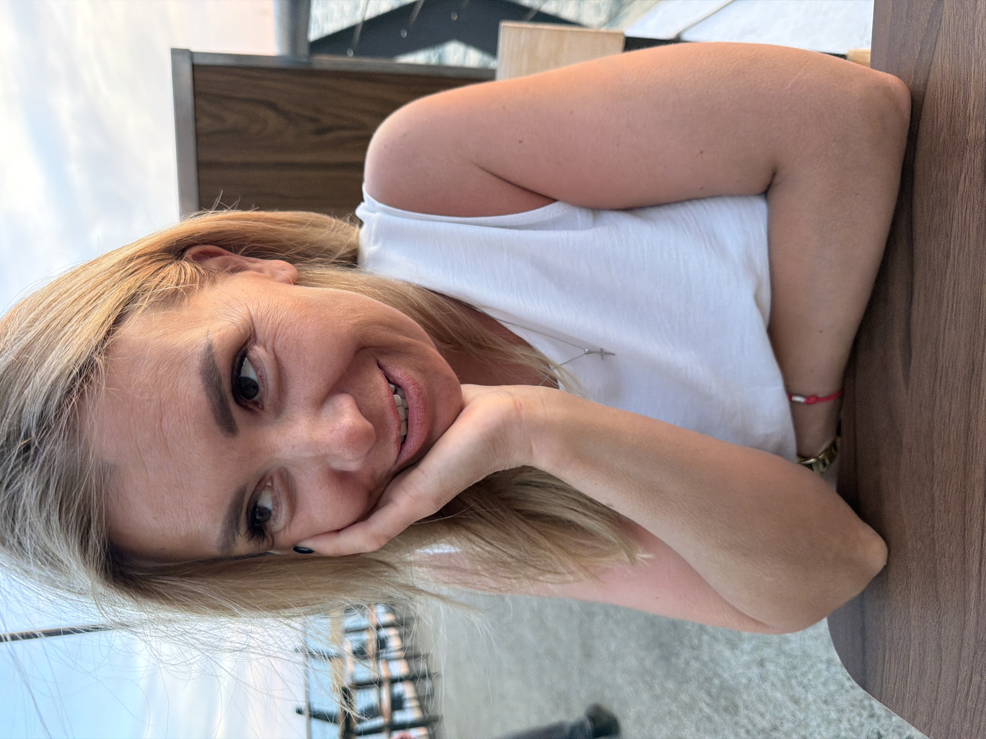 A woman with blonde hair is captured in a close-up, sideways portrait. She is resting her head on her hand, with her eyes looking towards the camera and a slight smile on her face. She is wearing a white tank top, and a thin silver necklace is visible. A red string bracelet is on her right wrist.
The background appears to be an outdoor setting, possibly a cafe or restaurant, with wooden paneling and a glimpse of a cloudy sky. The lighting suggests it is daytime, likely late afternoon or early evening. The woman's relaxed pose and smile convey a sense of casual ease and pleasantness. There is no visible text in the image.