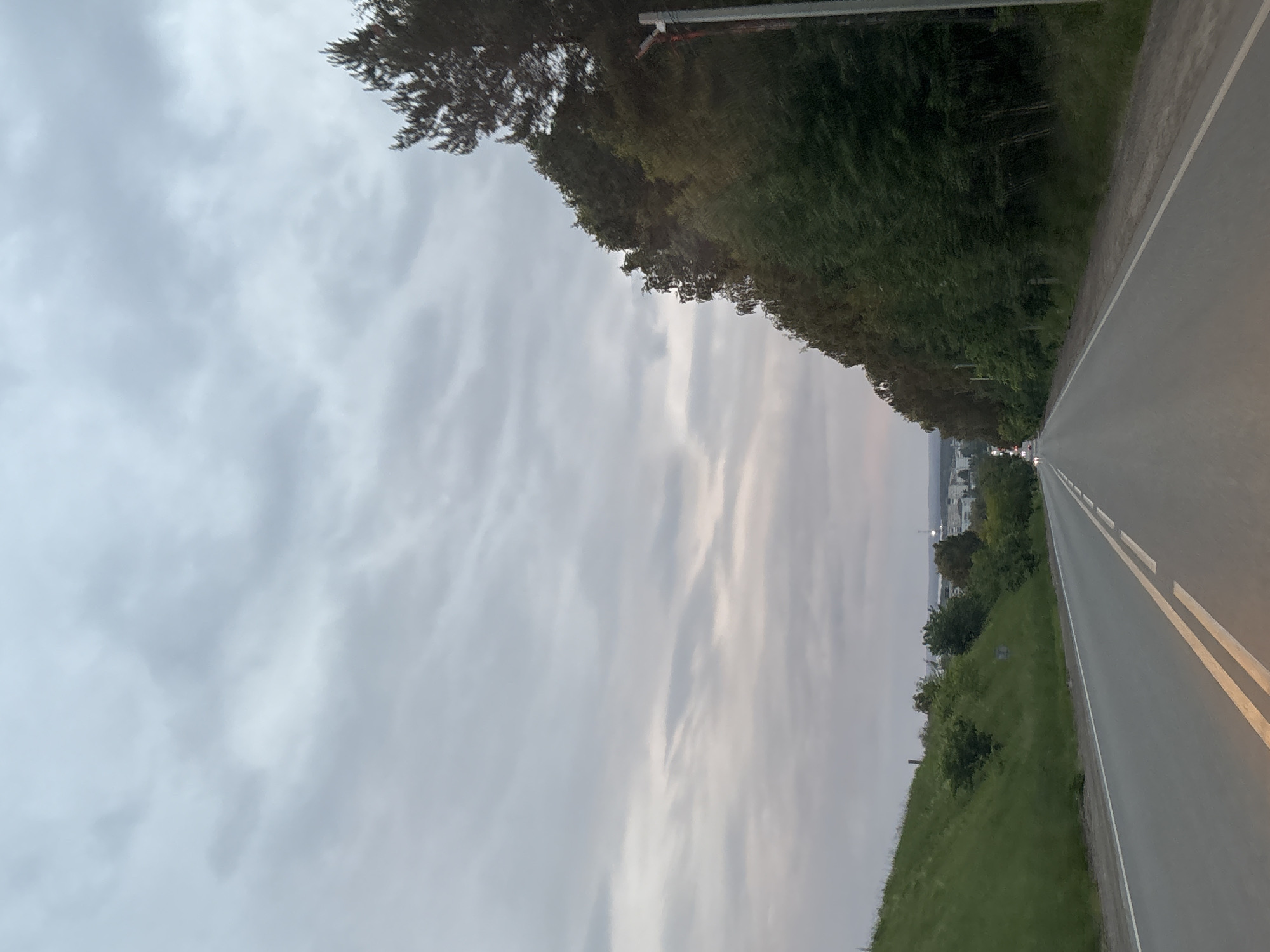 The image shows a road in Chaikovsky, Russia, with a cloudy sky. The road has two lanes in each direction and is bordered by a grassy hill on the left and trees on the right. In the distance, there are buildings and lights, suggesting an urban or suburban area. The lighting indicates it is likely dusk or early morning. There are no people visible in the image.