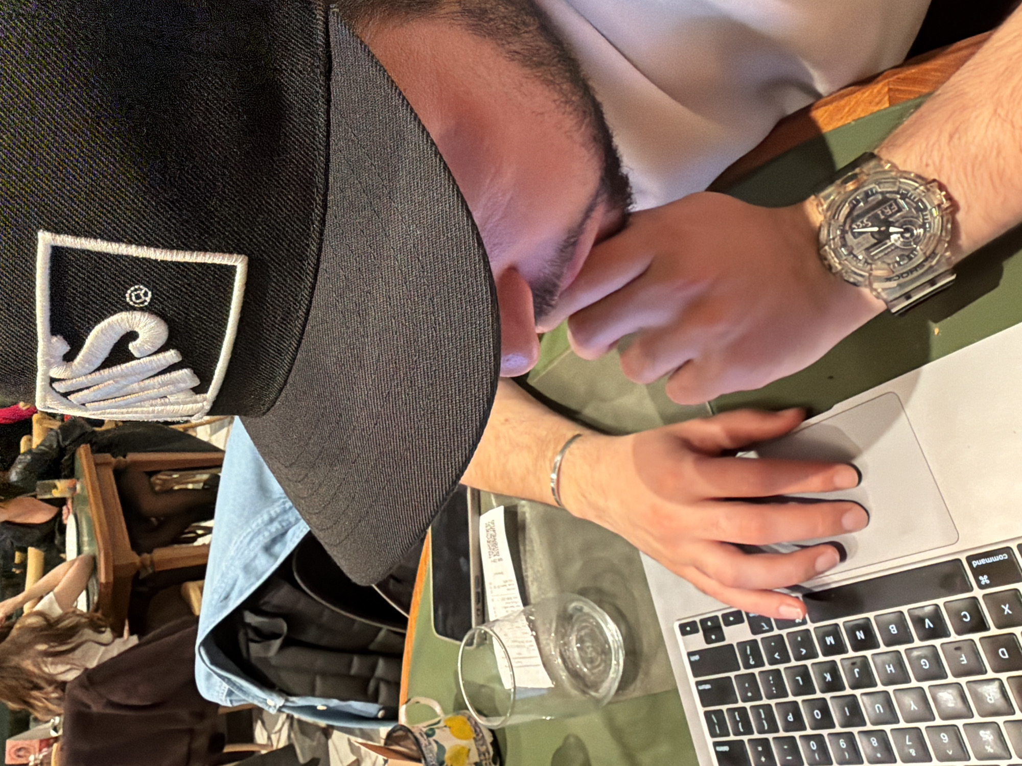 A man wearing a black cap with a white logo is seated at a table, operating a laptop computer. He has a clear wristwatch on his left wrist and a silver bracelet on his right wrist. There is a glass and a mug on the table near the laptop. The location is a public space, possibly a cafe or restaurant. The keyboard layout suggests it is in an English-speaking locale. There is a person sitting in the background. The setting is an interior space with artificial lighting. There is no indication of the location being Yerevan, Armenia.