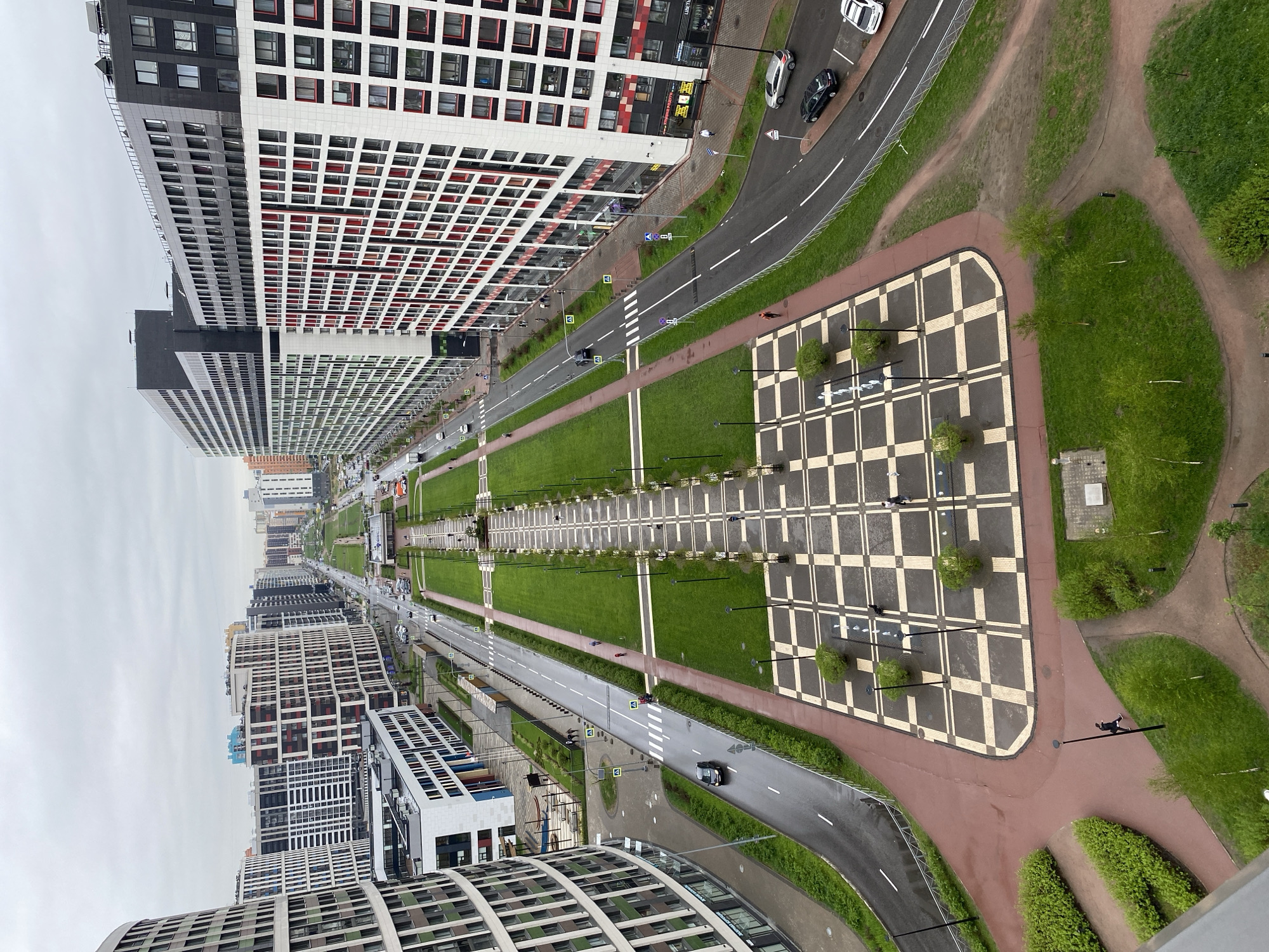 The image depicts an aerial view of a modern urban landscape in an unidentified city in Russia. The scene is dominated by contemporary high-rise apartment buildings with distinctive grid-like facades in shades of white, grey, and black, and occasional red accents. A long, narrow park stretches through the center of the scene, featuring manicured lawns, rows of trees, and decorative lighting. Adjacent to the park are roadways with moving vehicles and pedestrian walkways. A checkered-patterned plaza with trees and lighting fixtures is visible at the far end of the park. The weather appears overcast. Pedestrians can be seen walking on the pathways.