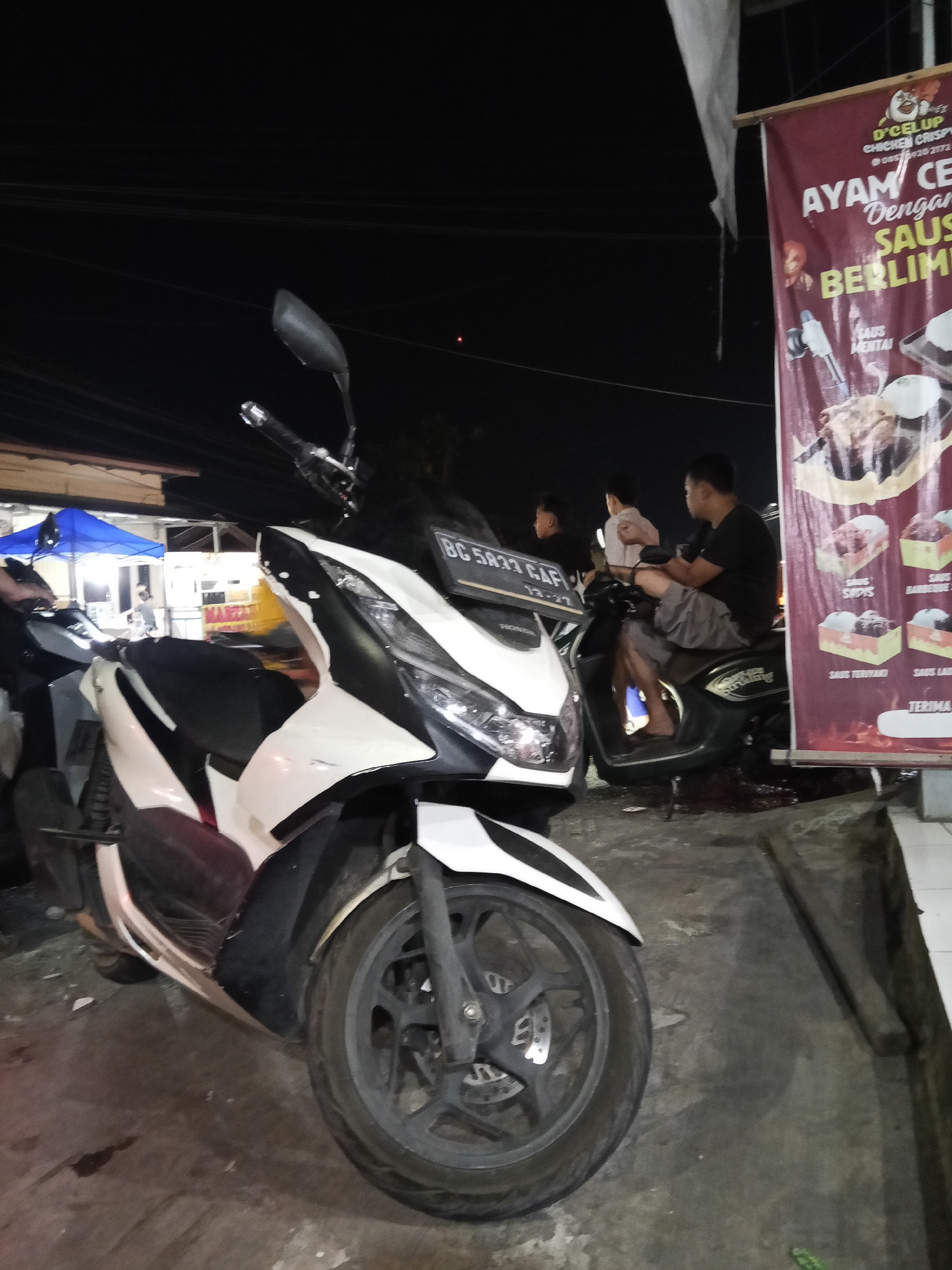 At night in Lubuklinggau, Indonesia, a white and black motorcycle is parked in the foreground on a dark, wet asphalt surface. Behind it, a dark green scooter carries three individuals. A man wearing a black t-shirt and grey shorts is seated on the scooter, holding the handlebars. To his left, a child in a light-colored shirt is also seated, and another young person with dark hair is positioned behind them. A banner with red and gold accents is visible to the right of the scooter. The banner features text in Indonesian and images of food, appearing to be an advertisement for a food stall. In the background, a blue tent is set up, illuminated by interior lights, suggesting a marketplace or food vendor area. The overall scene is dimly lit, typical of a street-side establishment operating after dark.