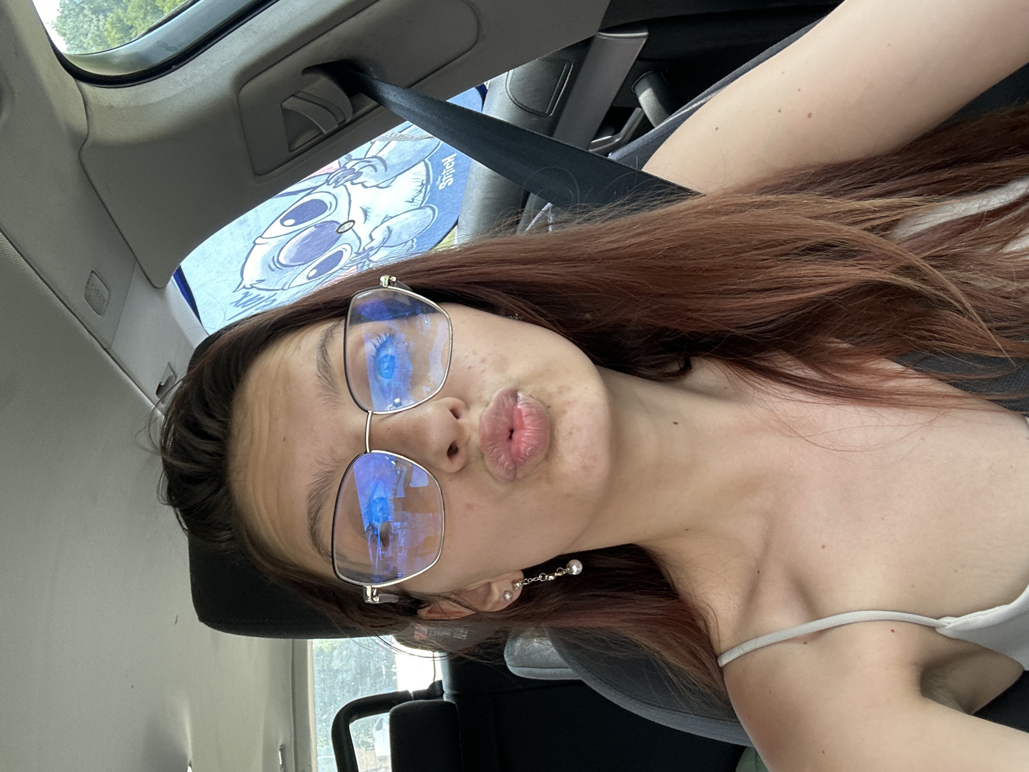 A young woman with long, reddish-brown hair and fair skin is sitting in the passenger seat of a car. She is wearing large, silver-rimmed glasses that reflect the blue sky. She has on a light-colored spaghetti-strap top and a dangling earring in her left ear. Her lips are pursed in a "kiss" pose. Behind her, a light blue sunshade with a cartoon character, possibly Stitch from Lilo & Stitch, is visible in the rear window. The interior of the car, including the headrest, seatbelt, and part of the dashboard, is visible. The image appears to be taken during the daytime, with natural light illuminating the scene. The location is identified as Tryavna, Bulgaria.