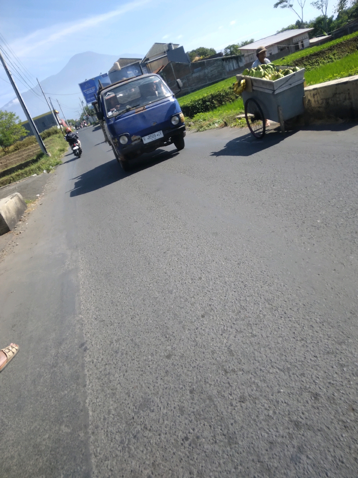The image depicts a street scene in Grendeng, Indonesia, on a clear day with a bright blue sky. In the foreground, the asphalt road shows signs of wear. To the left, a foot wearing a sandal is visible on the road's edge. A blue truck is driving on the road, with its license plate displaying "R 9509 CR". A person is riding a motorcycle further down the road, heading in the same direction as the truck. To the right of the road, a man wearing a hat is pushing a cart filled with produce, possibly bananas. The background features houses and agricultural fields, with mountains visible in the distance. Electrical poles and wires are also present along the roadside.