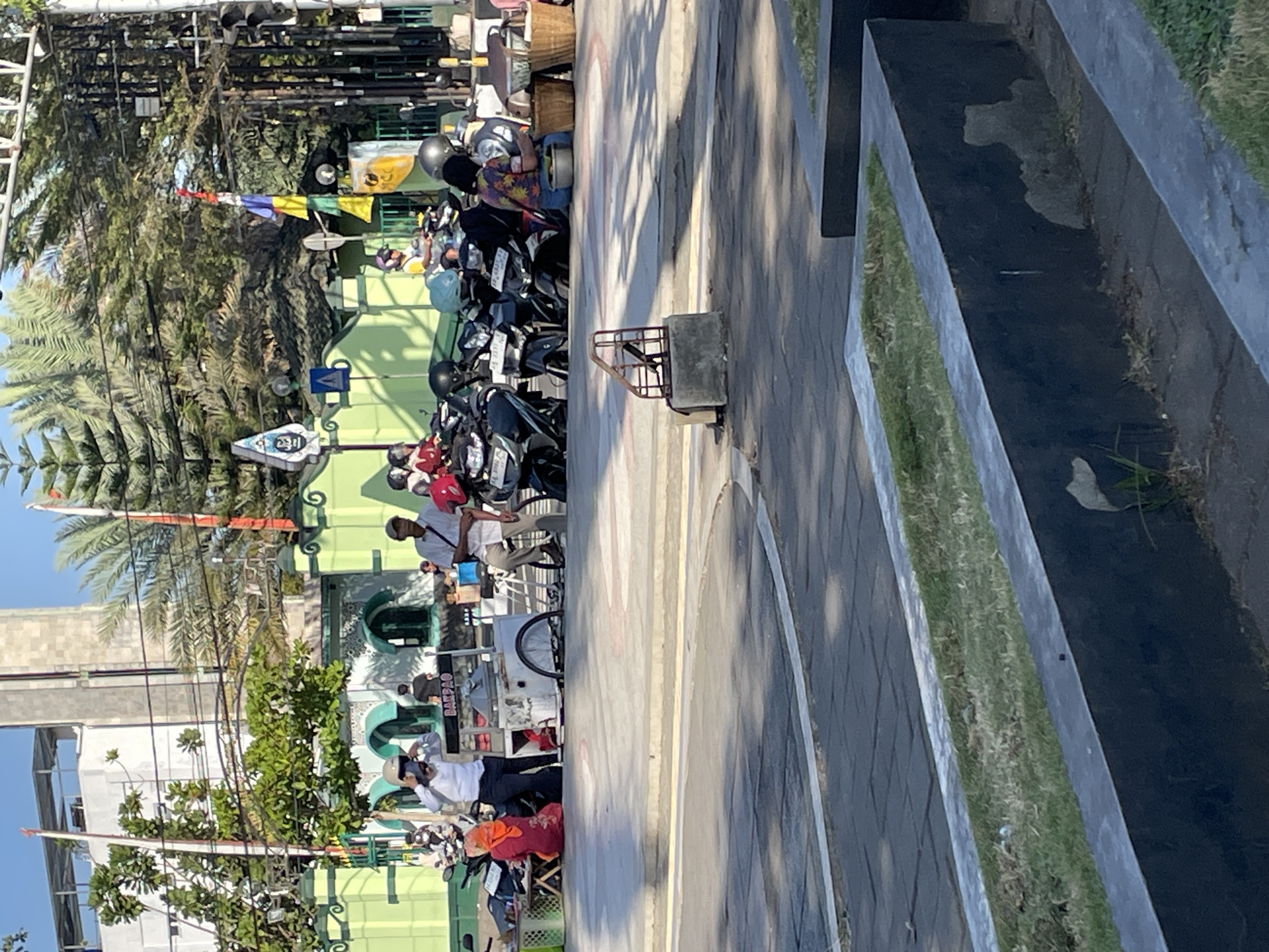 The image depicts a street scene in front of a mosque in Purworejo, Indonesia. In the foreground, there are several rows of black planters with grass. In the middle ground, parked motorcycles line the street, and several people are visible. A person in a white shirt and dark pants stands near a food cart labeled "BAKPAO." Another person in a red hijab and dress is seated nearby. A man in a white shirt and beige pants is also present. Behind them, the facade of a green mosque with intricate white patterns is visible, topped by a tall minaret. Palm trees and other foliage surround the mosque. The sky is clear and blue. The overall impression is a bustling daytime scene at a religious site.