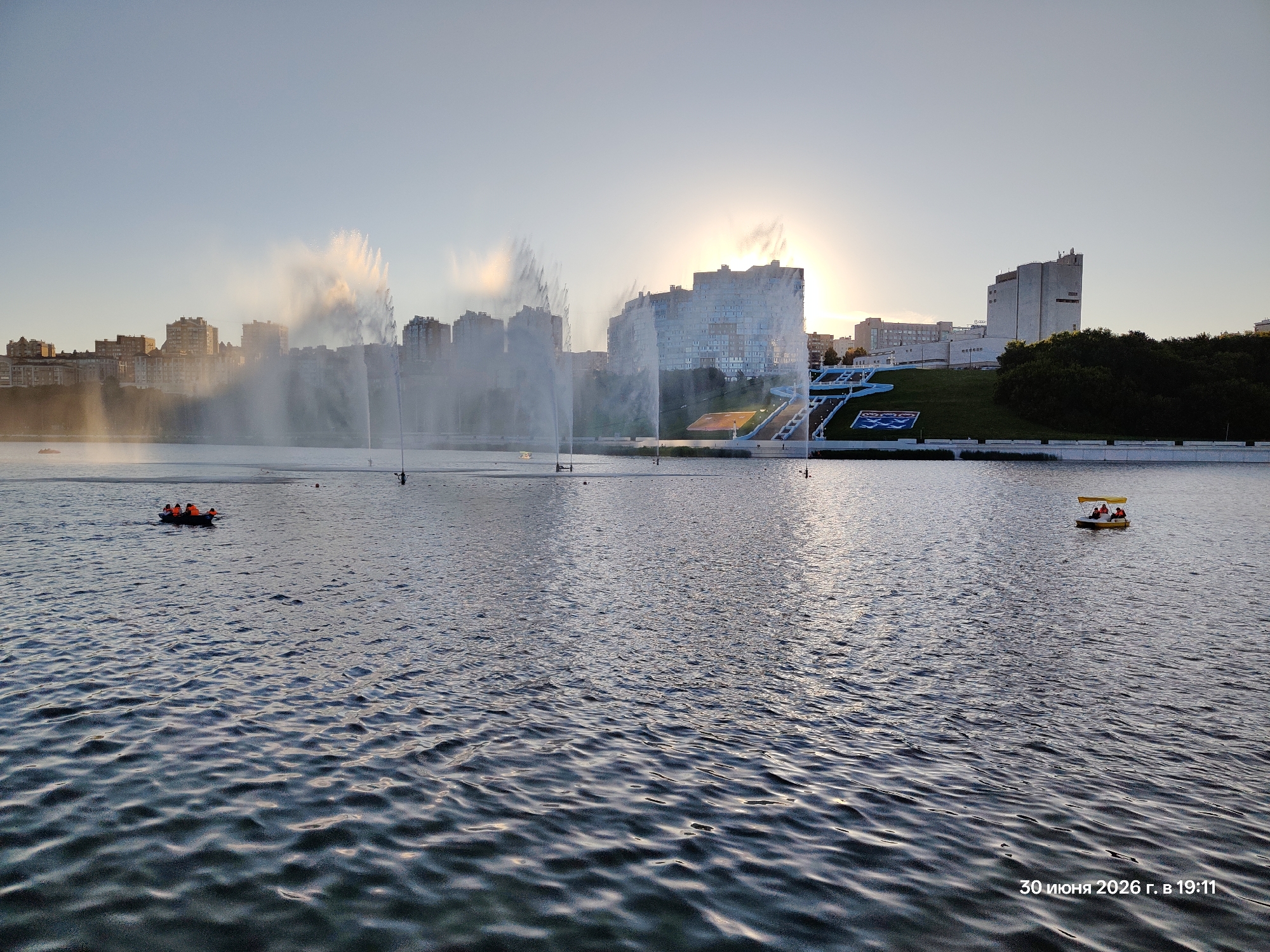 This media file captures a scenic view of a wide body of water with a cityscape in the background. Several tall buildings, some appearing residential and others more institutional, form the skyline. Large, dramatic fountains are active in the water, spraying plumes of water high into the air, catching the sunlight. The sun is low on the horizon, suggesting either sunrise or sunset, and its rays are partially obscured by the fountain mist, creating a soft, hazy glow.
On the water, several small boats are visible. One boat in the foreground left appears to be a paddleboat with a few people wearing life jackets, paddling. Further out, another small yellow boat with a canopy is also carrying people. These boats suggest recreational activity on the water. The water itself is rippled, reflecting the sky and the fountain spray.
In the background, there's a distinct tiered structure with white railings, possibly a promenade or viewing area, leading up to some buildings. Lush green trees line a section of the embankment. The overall atmosphere is serene and peaceful, with the soft light and the gentle motion of the water and fountains contributing to a tranquil mood.
Visible text in the bottom right corner reads "30 июня 2026 г. в 19:11", which translates to "June 30, 2026 at 19:11", indicating the date and time the image was taken. The weather appears clear with some atmospheric haze from the fountains.