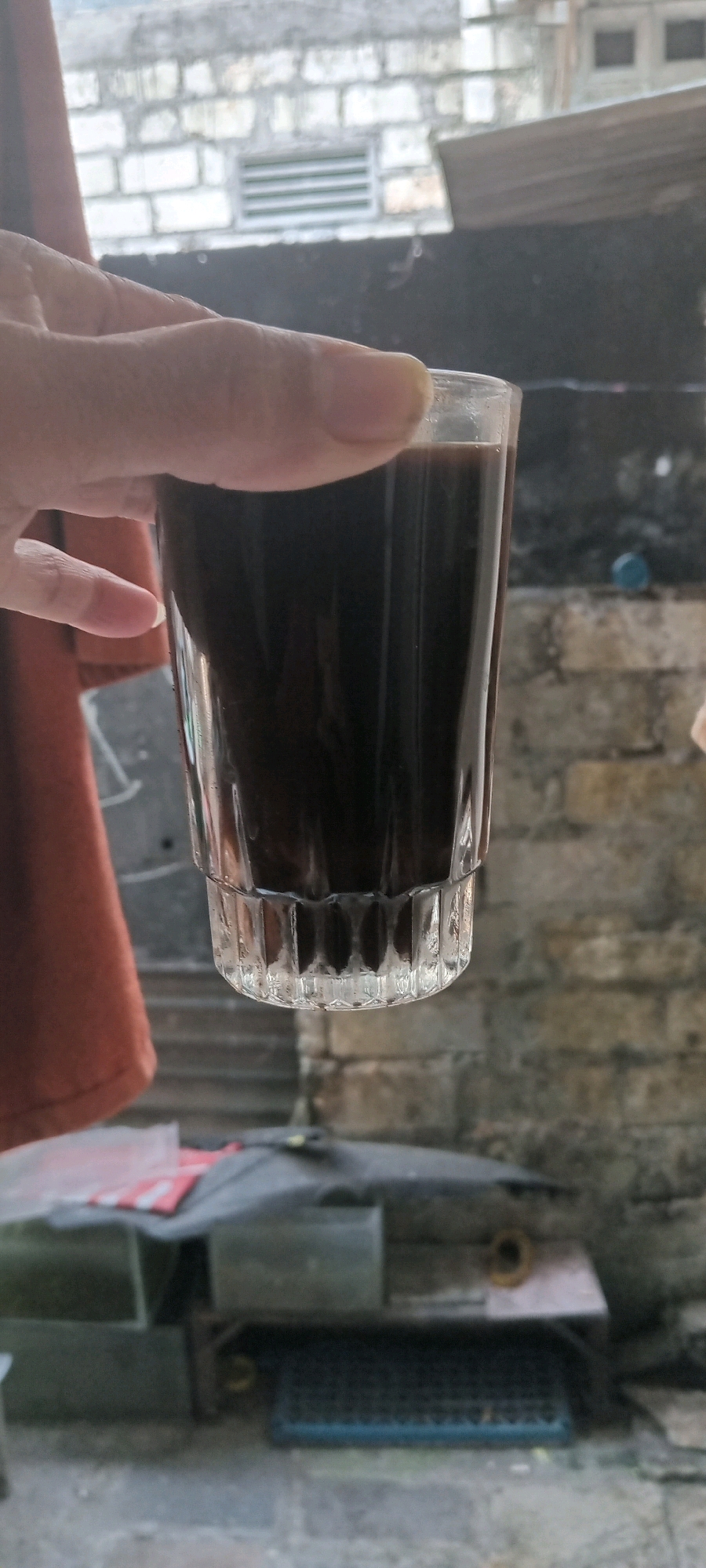 In Babat, Indonesia, a hand is holding a short, clear glass filled with dark liquid, likely coffee. The glass has a ribbed texture at its base. The background is slightly out of focus, showing a rough brick wall and some construction elements, possibly an outdoor area or alleyway. A reddish-brown cloth hangs on the left side of the frame. The lighting suggests it might be daytime, with a soft, diffused light.