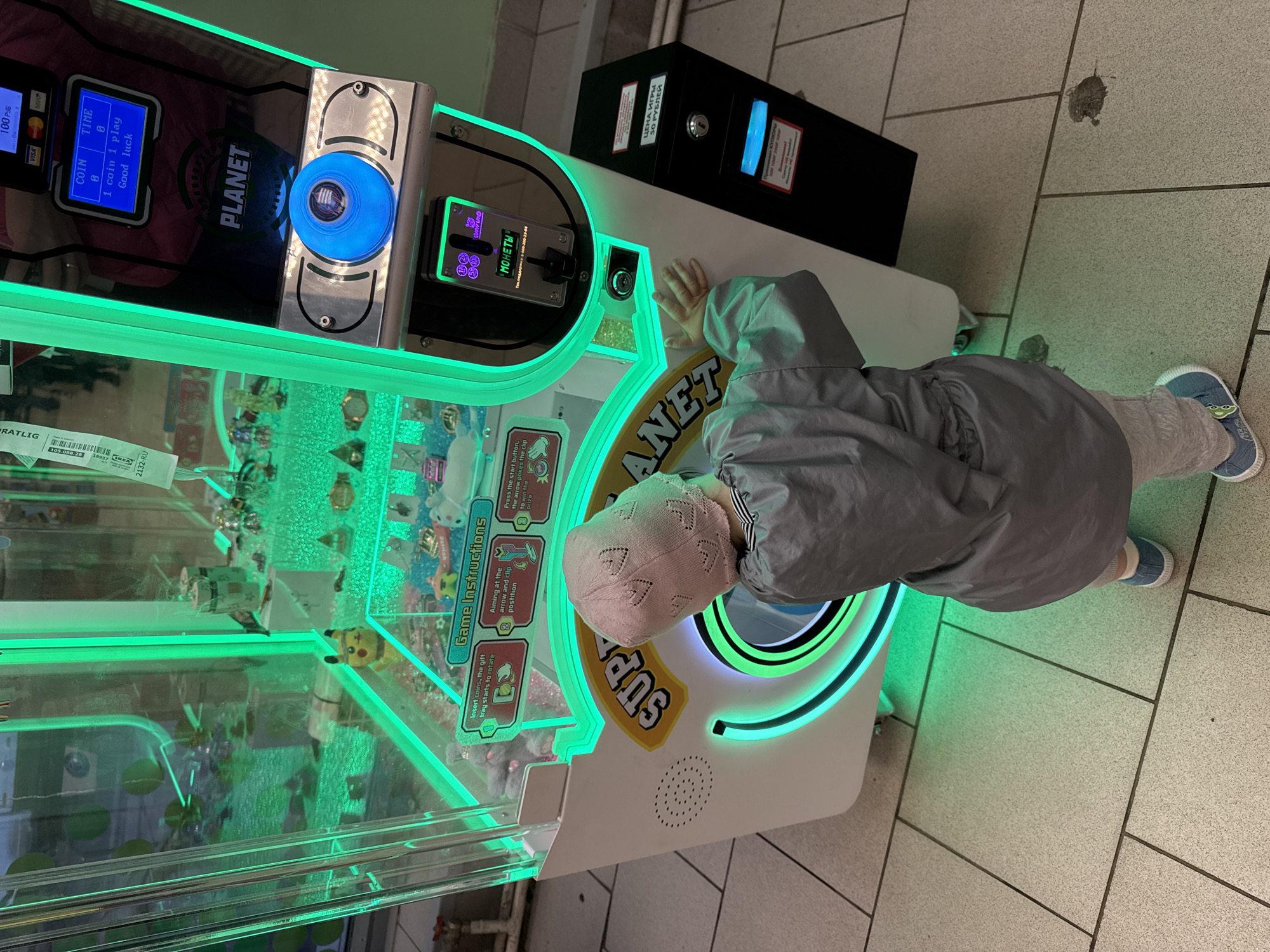 A child is playing a crane game at an arcade. The crane game has a green LED light outline, a blue start button, and a screen displaying the price of the game in rubles. There are game instructions on the machine. The game is called Planet Super. The child wears a grey hooded jacket, tan pants, a white knit hat with hearts, and blue shoes. The floor is tiled. Text on the crane game says "МОНЕТЫ" (coins), "ЦЕНА ИГРЫ 50 РУБЛЕЙ" (price of game 50 rubles), and the IKEA brand name "RATLIG" on a sticker. The arcade appears to be located in Moscow, Russia based on the language on the machine.