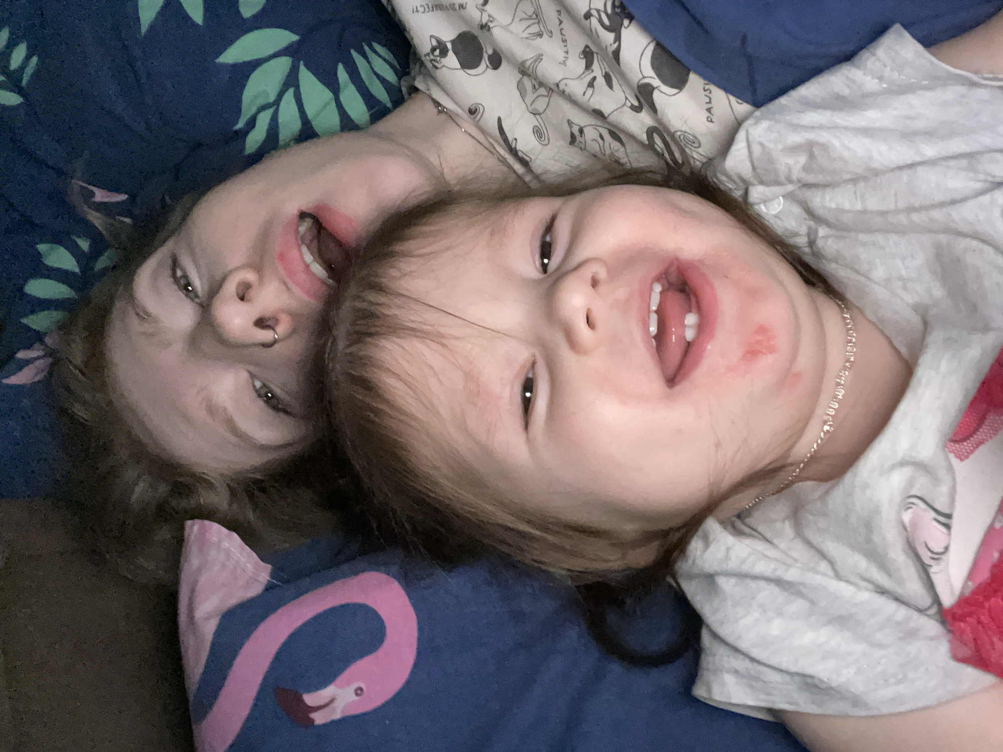 Two individuals, an adult and a child, are lying close together on a bed. The adult has brown hair and a nose ring, and is wearing what appears to be pajamas with a patterned top. The child, likely a toddler, is also wearing pajamas, and has a silver necklace. Both are smiling with their mouths open. The bed has a dark blue sheet with a pink flamingo design, and a green leaf pattern on a blue background is visible behind the adult. The location is not discernable from this image and is therefore not in Izhevsk, Russia.