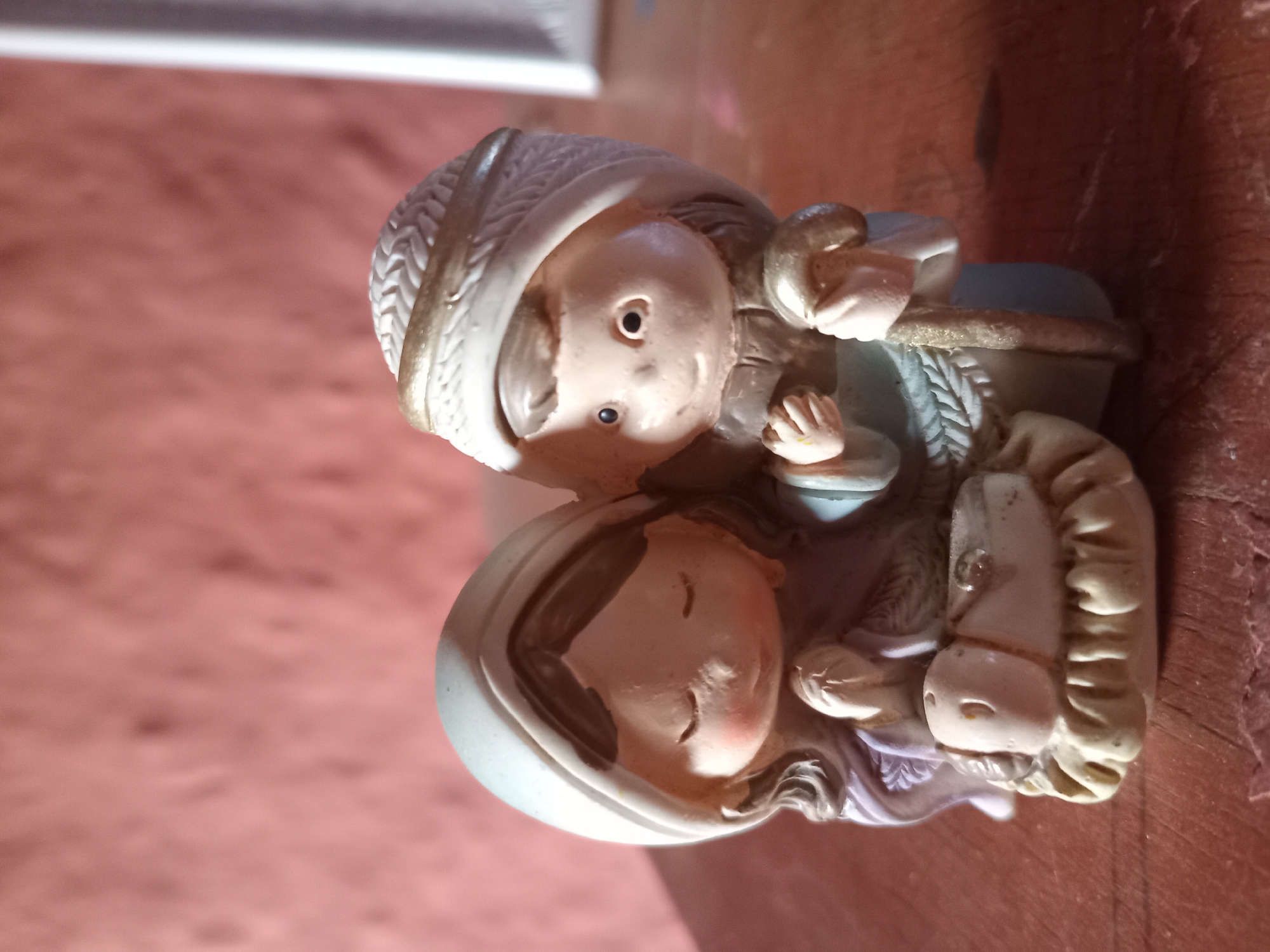 This is a close-up, eye-level photograph of a nativity scene figurine set on a wooden surface. The figurine depicts Mary, Joseph, and the infant Jesus in a manger. Mary is on the left, wearing a blue head covering and a purple garment, with her hands clasped in prayer. Joseph is on the right, wearing a white head covering and a blue tunic, holding a shepherd's staff. The infant Jesus is in a manger in front of them. The background is a textured, reddish-pink wall. The scene appears to be indoors, likely in a domestic setting in Juan Griego, Venezuela, though specific landmarks are not visible.