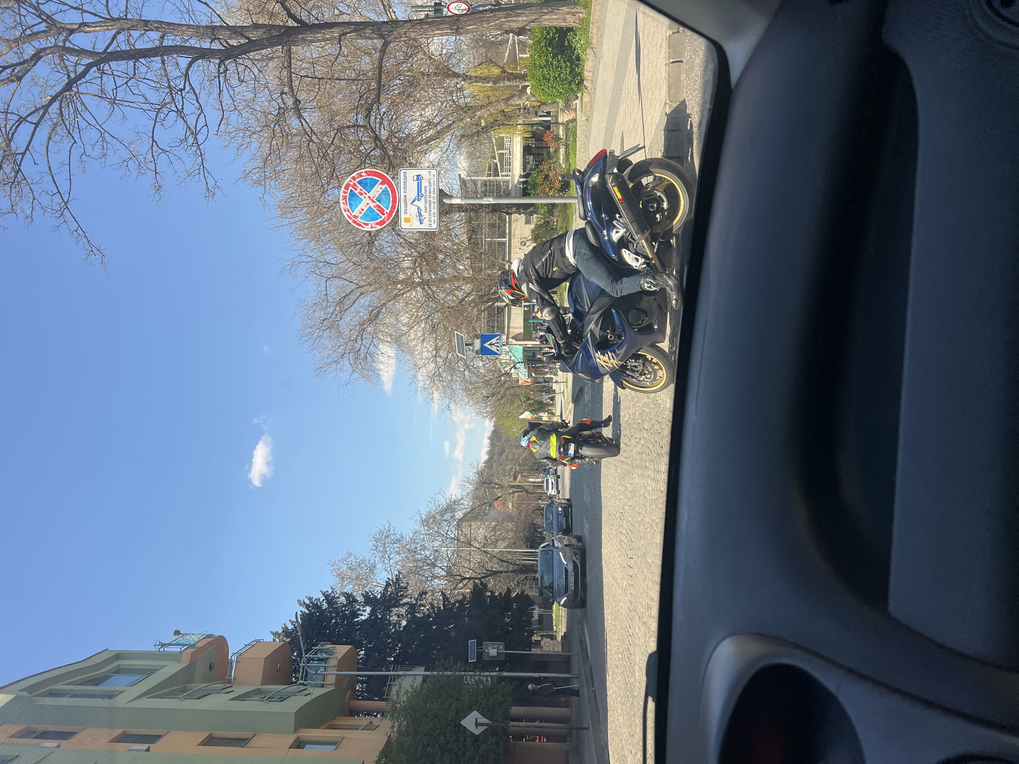 The image shows a street scene in Plovdiv, Bulgaria, likely taken from inside a parked car. The day is sunny with a clear blue sky. A "no stopping" sign is visible on the right side of the street. Several motorcyclists are parked on the sidewalk next to the sign. Behind them, cars are parked along the street. Buildings and trees line the left side of the road, and hills are visible in the distance.
