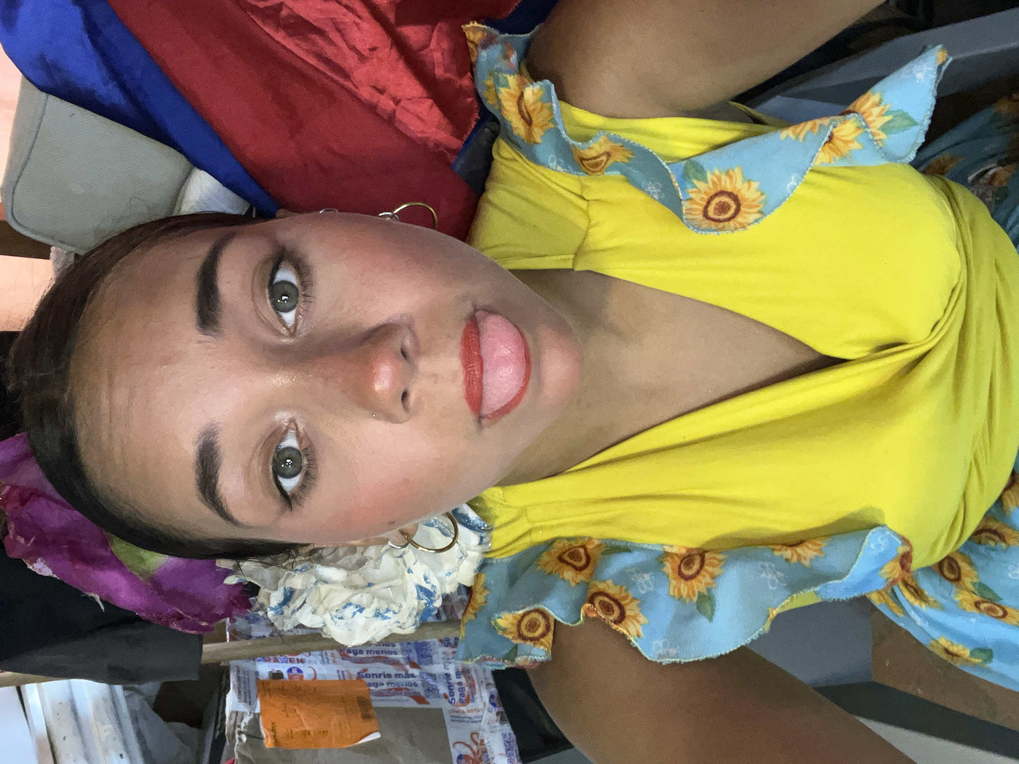 A young woman is prominently featured in a close-up, head-and-shoulders shot, tilted to the side. She has dark hair, green/hazel eyes, and well-defined eyebrows. Her face shows some redness on the cheeks, possibly from blush, and she is wearing red lipstick. She is playfully sticking out her tongue towards the viewer. A gold hoop earring is visible in her right ear (viewer's left).
She is wearing a bright yellow top with ruffled sleeves that are light blue and patterned with yellow sunflowers. The background behind her upper body consists of draped fabrics in red and blue, along with a light grey object that might be a cushion or part of a bag. Near her head, to the left, there is a cluster of white, crumpled material that resembles paper flowers or decorations, with hints of purple fabric mixed in. Below this, there are various items, including crumpled paper or packaging.
Visible text on the crumpled paper in the lower part of the image includes phrases in Spanish: "Sonrie mas Paga menos" (Smile more Pay less), "Paga menos" (Pay less) repeated, and partially visible words like "Ahorra" (Save) and "Sonrisa" (Smile). The overall impression is informal and candid, with a cheerful and playful mood, possibly captured indoors in Porlamar, Venezuela.