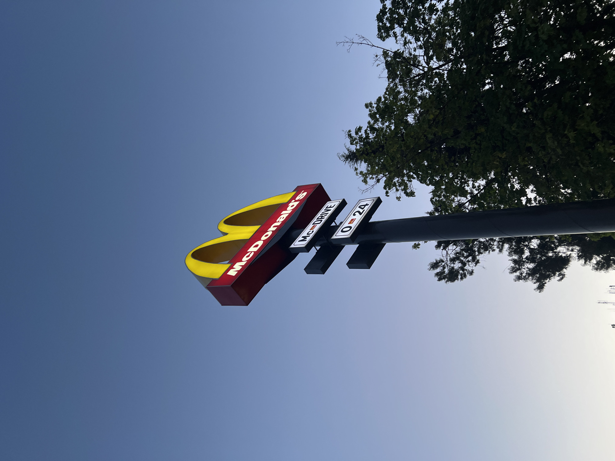 This upward-angled photograph captures a McDonald's sign set against a gradient blue sky, with a large tree partially visible on the right. The prominent golden arches of the McDonald's logo are visible, with the red rectangular base displaying the illuminated white text "McDonald's".
Below the main sign, two smaller, rectangular white signs are attached to a dark vertical pole that supports the entire structure. The upper small sign clearly reads "McDRIVE", indicating the presence of a drive-through service. The lower small sign displays "0-24", suggesting the establishment operates 24 hours a day.
To the right side of the frame, a large tree with dense green foliage is visible, its branches and leaves silhouetted against the bright sky. The sky itself transitions from a deep, rich blue at the top and left to a lighter, almost whitish-blue towards the bottom and right, indicative of a clear day with the sun likely low on the horizon, possibly morning or late afternoon. There are no clouds, and the weather appears calm and bright. No people or activities are present in the image, focusing solely on the static sign and natural elements. The scene is located in Plovdiv, Bulgaria.