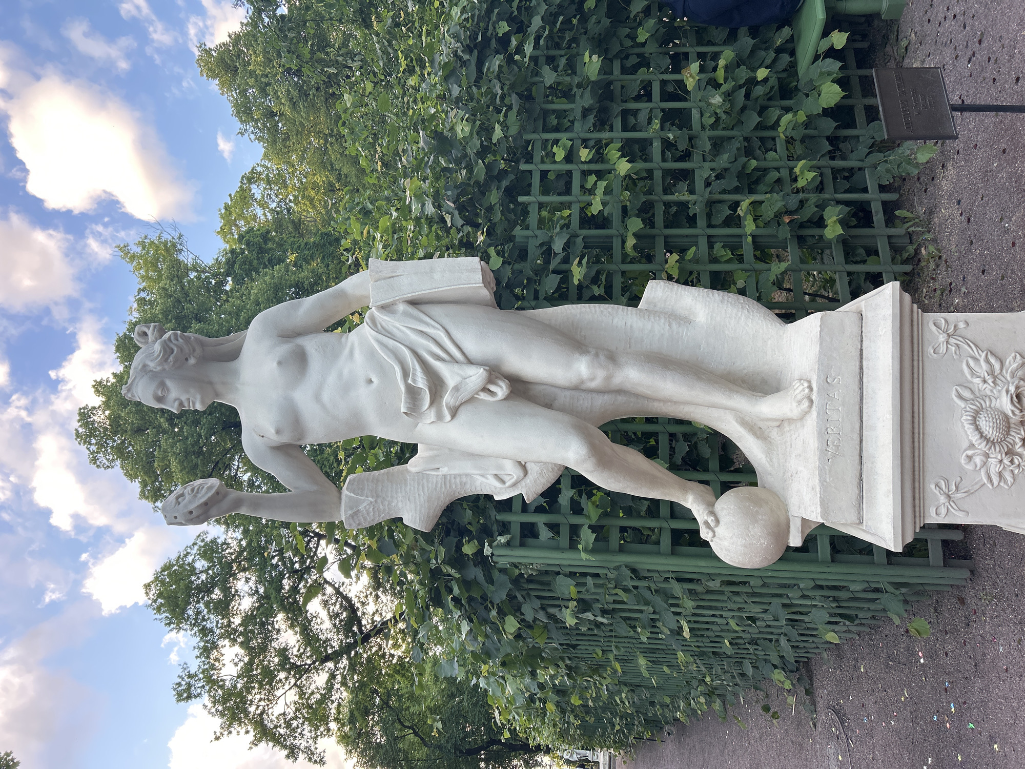 An eye-level, outdoor, full shot captures a white marble statue of a male figure on a pedestal in a park. The statue, which stands on a square base, is positioned on the right side of the frame, with its head turned to the left. The figure is nude and appears to be in motion, with its left leg bent and forward. In its right hand, the figure holds a circular object, and in its left hand, it holds a rectangular object. The pedestal is decorated with carvings of flowers. The background is a lush green hedge, behind which trees can be seen. The sky above is a light blue with scattered white clouds. A dark green metal fence runs along the left side of the statue and extends into the background. A paved walkway is visible to the left of the fence. In the lower right corner, a dark sign with text is planted in the ground. The overall scene is peaceful and serene.