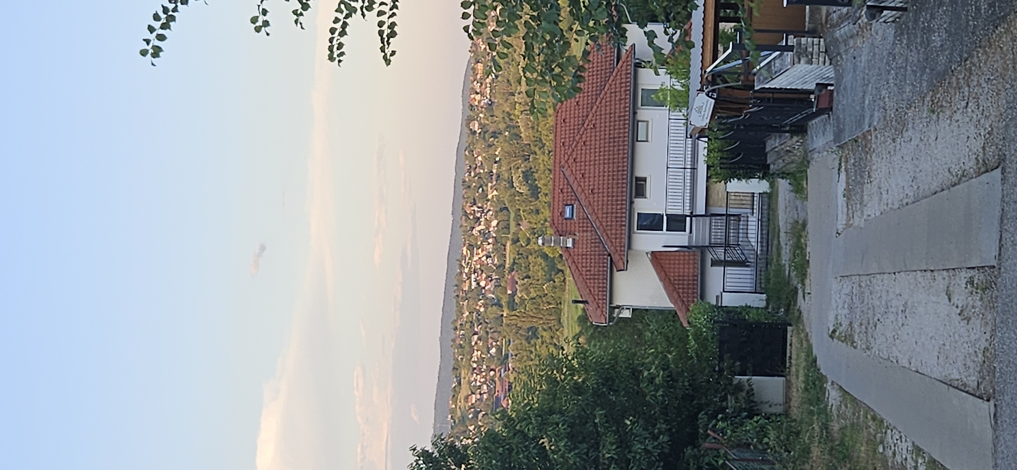 The image depicts a hillside residential area in Hévíz, Hungary, bathed in the soft light of late afternoon or early evening. The foreground features a concrete driveway or pathway leading towards a multi-story house with a red tiled roof. This house has white walls, a balcony with black railings, and several windows. Adjacent to the house, there's a black metal gate and fencing, along with some greenery.
Behind the house, a vast expanse of houses and trees stretches across the undulating landscape, indicative of a suburban or rural setting within the city. The distant hills are visible on the horizon, under a pale blue sky with scattered, wispy clouds. The overall impression is one of a tranquil, settled community nestled in a verdant environment. There are no people visible in the image.