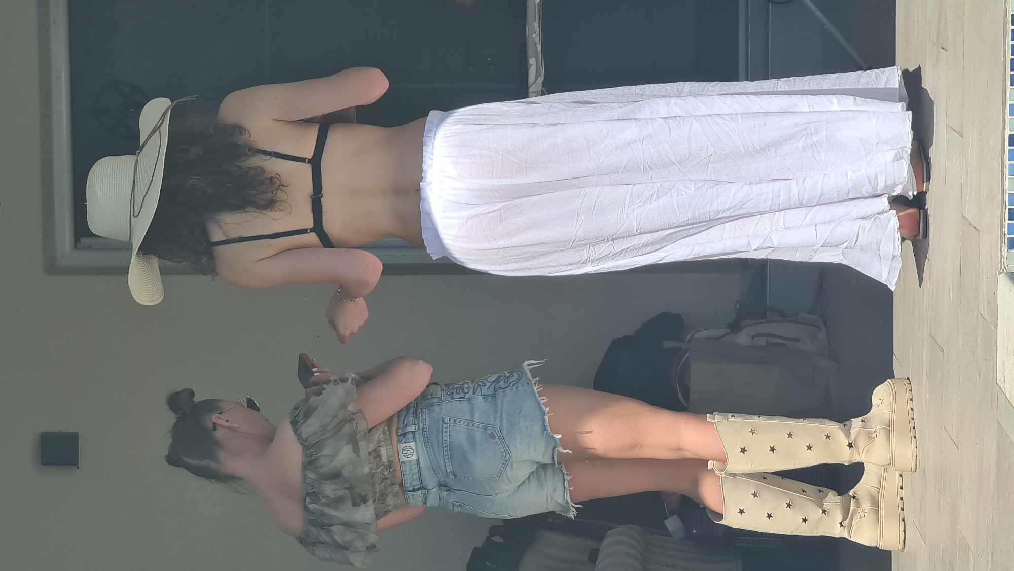 A photo taken at Dobra Voda, Montenegro, shows two women standing near a pool. The woman at the top is wearing a white sun hat, a black bikini top, and a long white skirt. The woman at the bottom is wearing sunglasses, a patterned top, denim shorts, and tall boots with star patterns.
