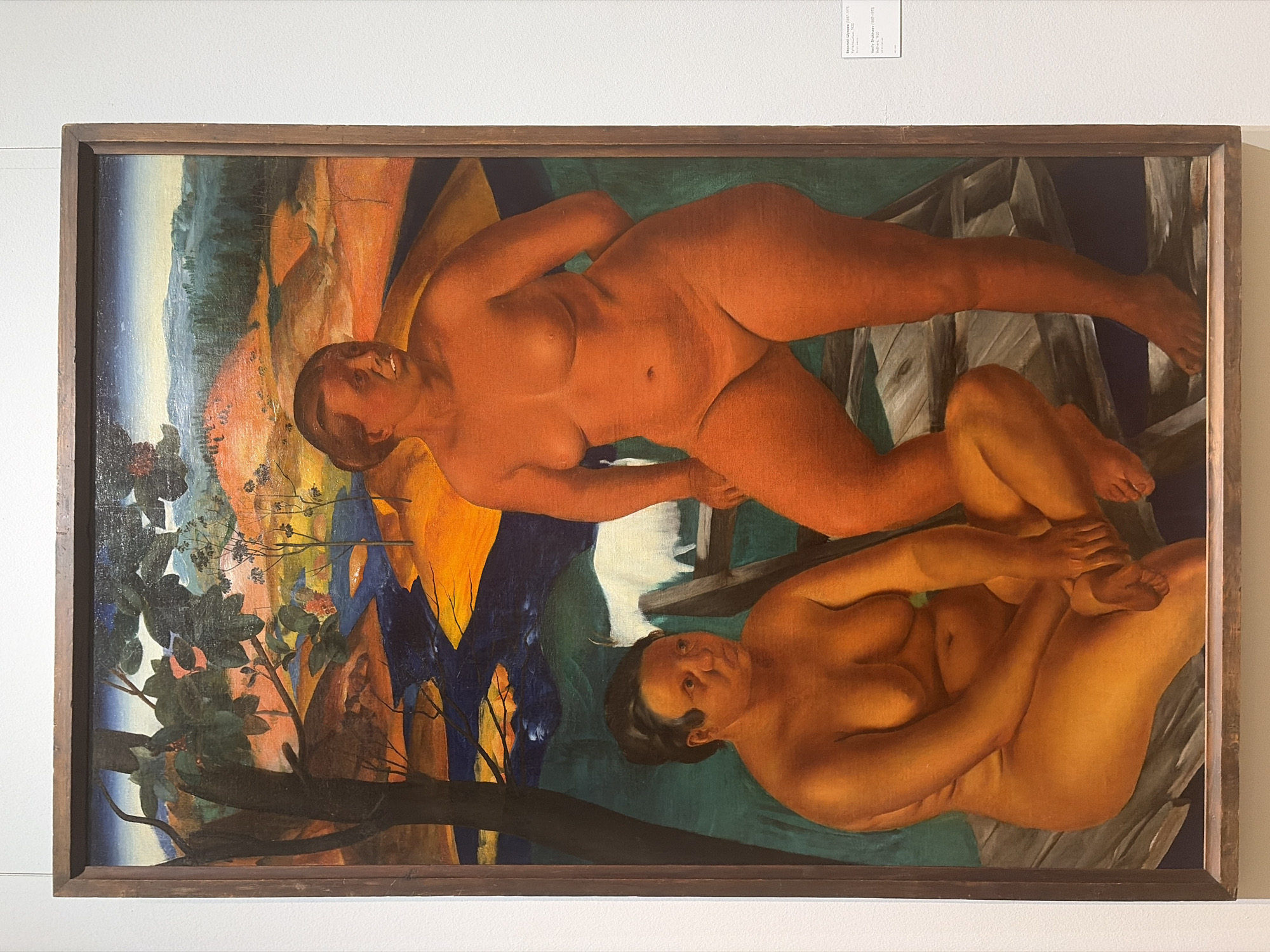 This is a painting titled "Bathers" by Vasily Shukhaev, created in 1920. The artwork depicts two nude female figures reclining in a natural setting. The figures are rendered in warm, earthy tones, with the upper figure in a reddish-brown hue and the lower figure in a yellowish-ochre. They are positioned in a way that suggests a moment of rest or repose.
The background is a vibrant and abstract landscape. To the left, there are stylized trees with dark green foliage, set against a backdrop of warm oranges, yellows, and blues that suggest hills or mountains under a dramatic sky. To the right, there are hints of wooden structures, possibly a bridge or a fence, rendered in a simplified, geometric style. The overall color palette is bold and expressive, with strong contrasts between light and shadow.
The painting is framed by a simple wooden border. A small informational plaque is visible above the painting, providing the title in both Russian and English, along with the artist's name and birth/death dates. The plaque also indicates that the artwork was created in 1920. The scene appears to be outdoors, possibly near water, given the title "Bathers," although the specific location is not explicitly detailed beyond being a natural landscape. The figures themselves do not display overt emotions, but their relaxed poses contribute to a sense of tranquility and naturalism.