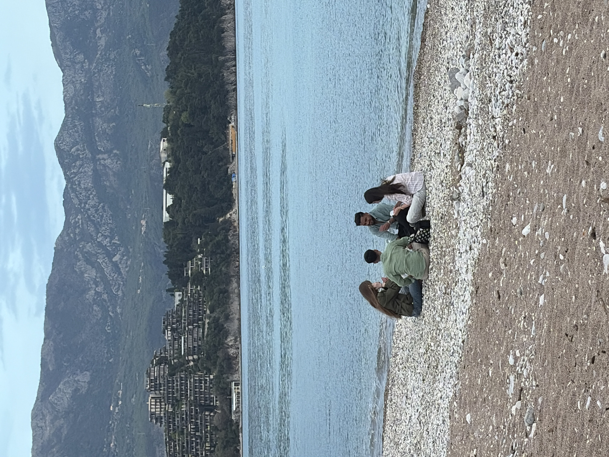 The image shows a group of four people sitting on a pebble beach next to calm blue water. Behind them are a hillside with buildings and tall mountains, partially obscured by a hazy sky. The scene is likely in Budva, Montenegro, given its coastal landscape and architectural style. The people appear to be engaged in a casual gathering, possibly conversing or enjoying the view. The beach surface transitions from pebbles near the water to a mix of sand and pebbles further inland.