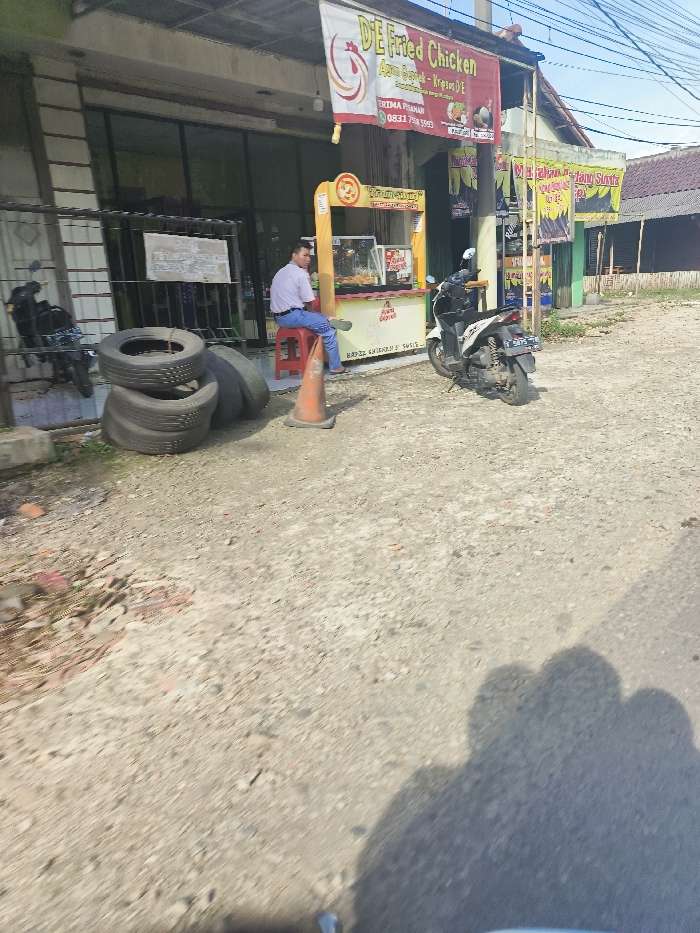 The scene is a street in Jatiluhur, Indonesia, with a small food stall on the right side. A man is sitting on a stool in front of the food stall, which is painted yellow and has a red awning. There is a motorcycle parked next to the stall, facing the street, with a red license plate. The man is wearing a light blue shirt and dark blue pants. There are multiple tires stacked in the background, along with a scooter and a red and white traffic cone. The street is paved with gravel and dirt. There are several buildings on the left side of the street. The food stall has a sign that reads "DE Fried Chicken" in English. The sign above it reads "Ayah Gagal - Krazy DE" in Indonesian. The street is relatively empty, with no other people visible. The sky is clear with light clouds and the sun is shining.