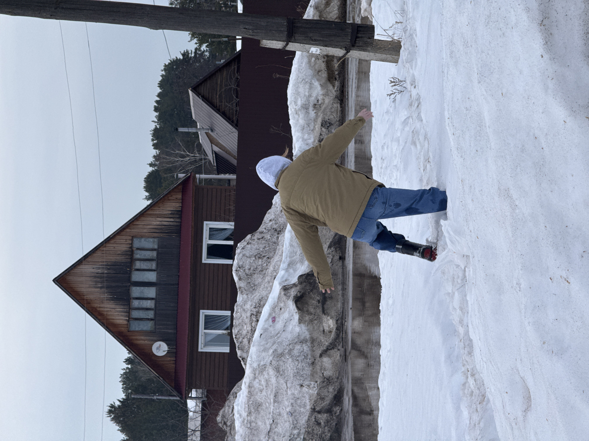 The scene captures a person navigating a thawing, snowy landscape with standing water in the city of Chaikovsky, Russia. An individual, viewed from behind, is positioned centrally, leaning forward with arms outstretched for balance. They wear a light-colored hooded sweatshirt, a brown jacket, and blue jeans, with the lower part of their left leg and a dark boot (featuring a red sole) submerged in shallow water or slush. The right leg is partially obscured by snow.
In the midground, a wide expanse of dark, still water reflects the overcast sky, bordered by large, irregular piles of dirty, melting snow. Behind these snow banks, a dark brown wooden fence runs horizontally. A two-story house with a dark brown lower facade and a natural wood gable roof with dark red trim is prominent in the background. It features two white-framed windows with curtains on the lower level and four smaller rectangular windows in the upper gable, along with a white satellite dish. To the right of this, another house with a dark roof is partially visible.
A dense line of coniferous trees forms the distant background under an overcast, light gray sky. A dark utility pole, reinforced with a concrete section and marked with a white sticker (partially illegible, possibly "1998"), stands on the far right of the frame, with overhead wires visible. The ground in the foreground consists of uneven, partially melted snow, with a small, dry shrub-like plant visible to the right of the person. The overall environment indicates a period of advanced thaw, likely late winter or early spring.