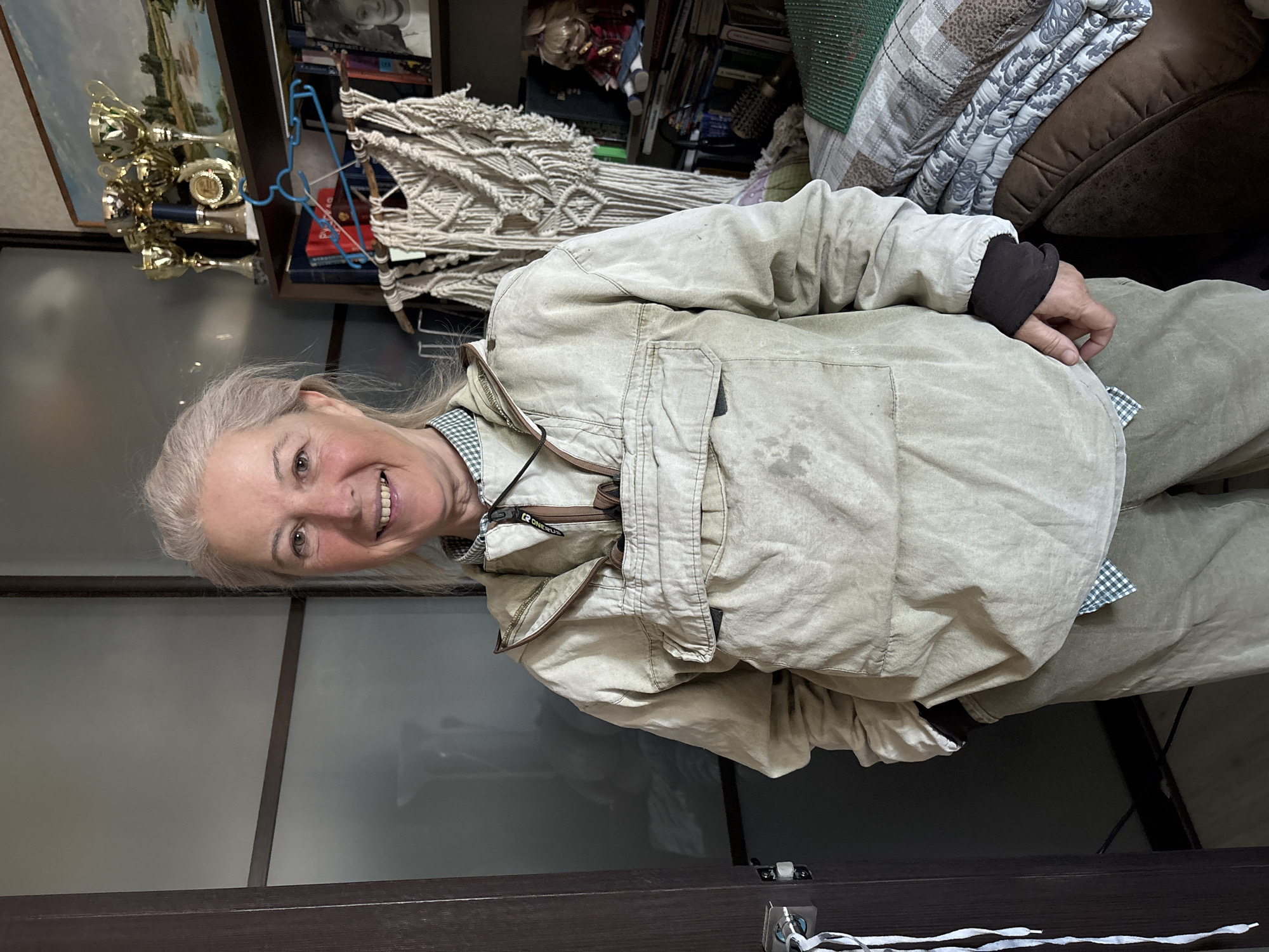 A woman with gray hair smiles at the camera. She is wearing a tan jacket and trousers. The background includes a shelf filled with trophies, books, and decorative items. The woman is indoors. The location is Ulyanovsk, Russia.