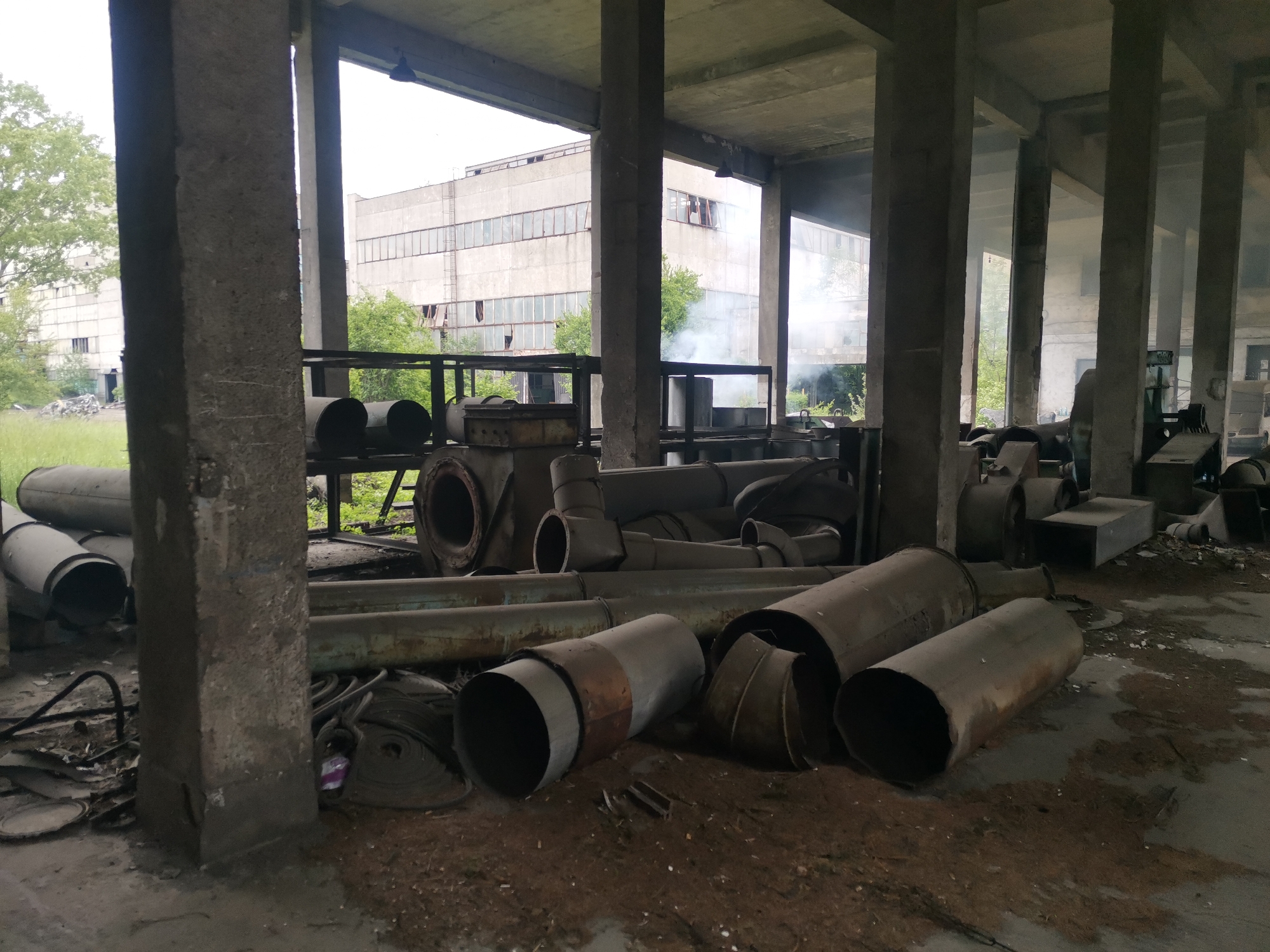 The image depicts an abandoned industrial site, possibly in Zhivkovo, Bulgaria, given the request context. The location features a large, concrete-columned building with a decaying interior. Various metal pipes and industrial equipment are scattered across the ground, amidst debris. Outside the structure, overgrown vegetation and another building can be seen. Smoke is visible in the background. No people are clearly visible in the image, but a person is off in the distant background.
