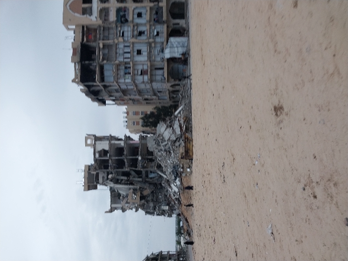 War-torn buildings are shown from a low angle, displaying extensive damage with crumbled structures and exposed interiors. Debris is scattered around the base of the buildings. The area appears barren, with a sandy or dusty surface in the foreground. Overcast skies create a somber atmosphere, highlighting the devastation and desolation of the scene.