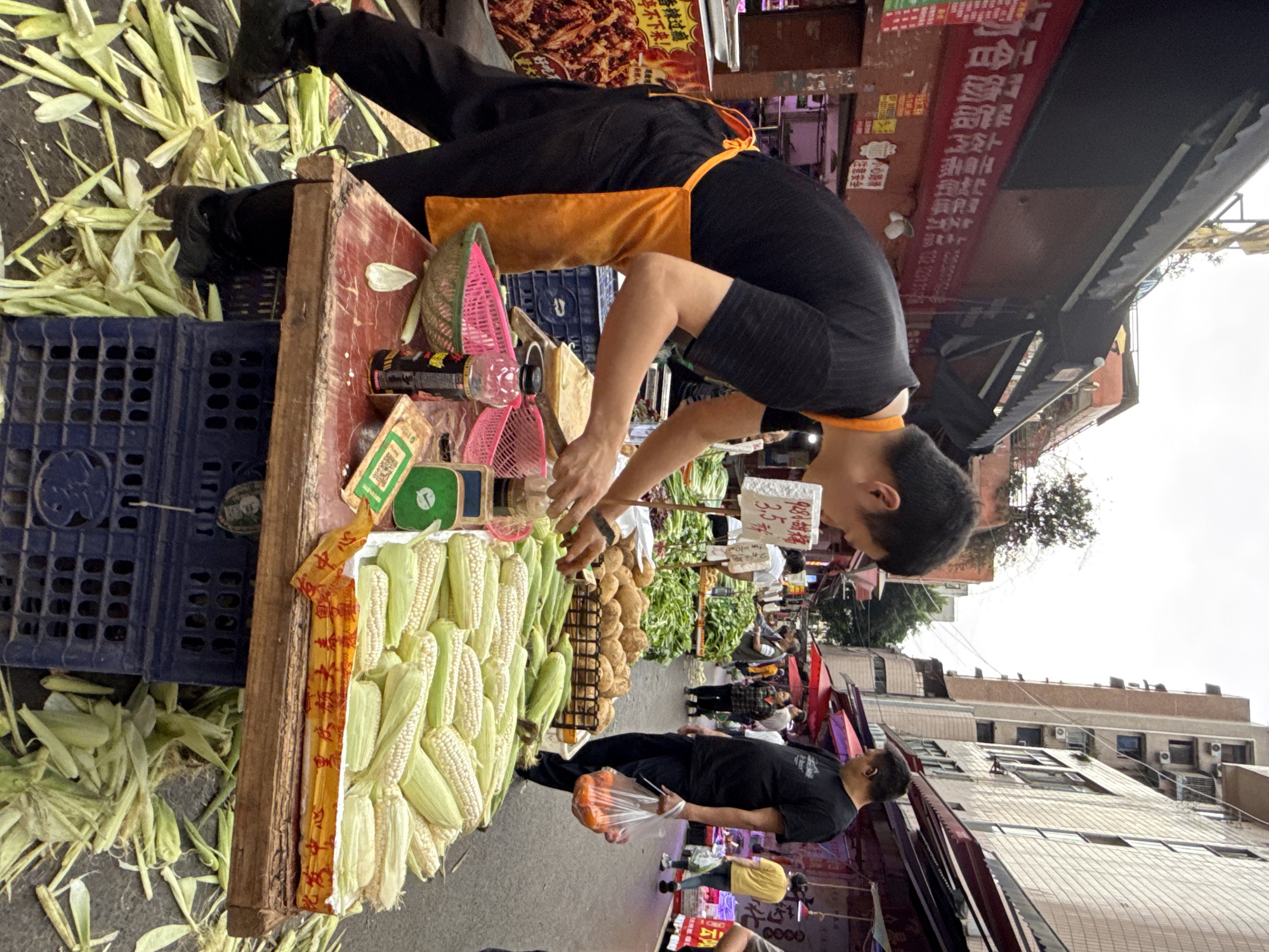 A street vendor is selling corn from a wooden table on a Nan'an District, China street. A blue crate sits beside the table, and corn husks litter the ground nearby. The vendor, wearing a black shirt and orange apron, is sorting through the corn. Customers are milling around the table, and other vendors are visible in the background. Buildings line the street. Signs with Chinese characters are visible.